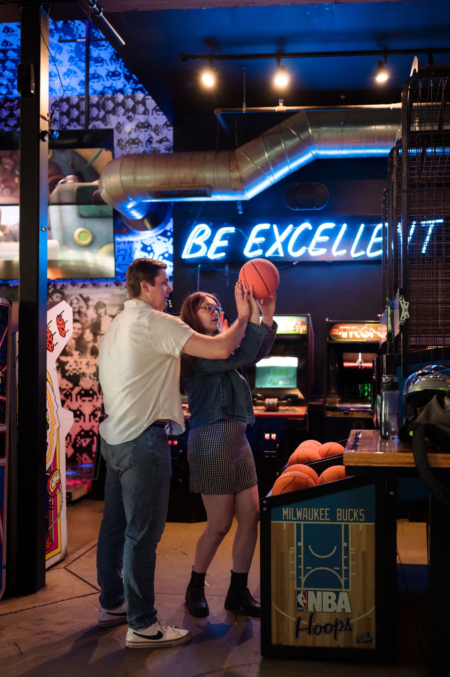 Couple playfully reaching for a basketball together inside a retro arcade bar with neon signs.