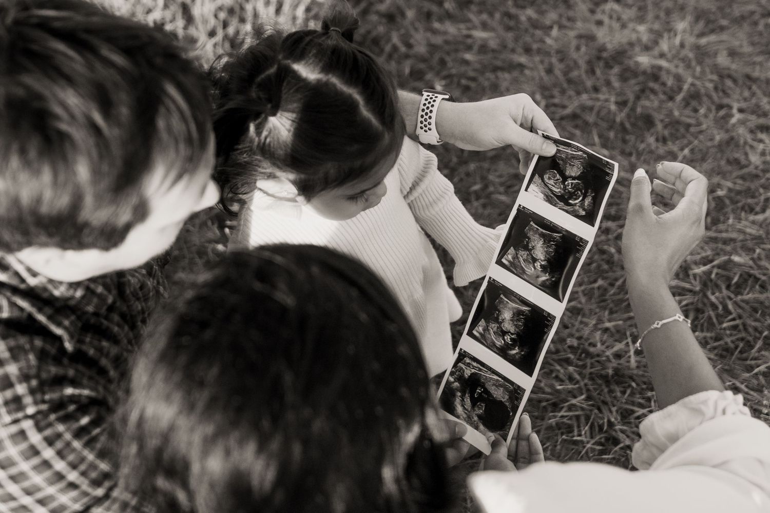 Family viewing ultrasound photos together on grass in black and white maternity announcement photo.
