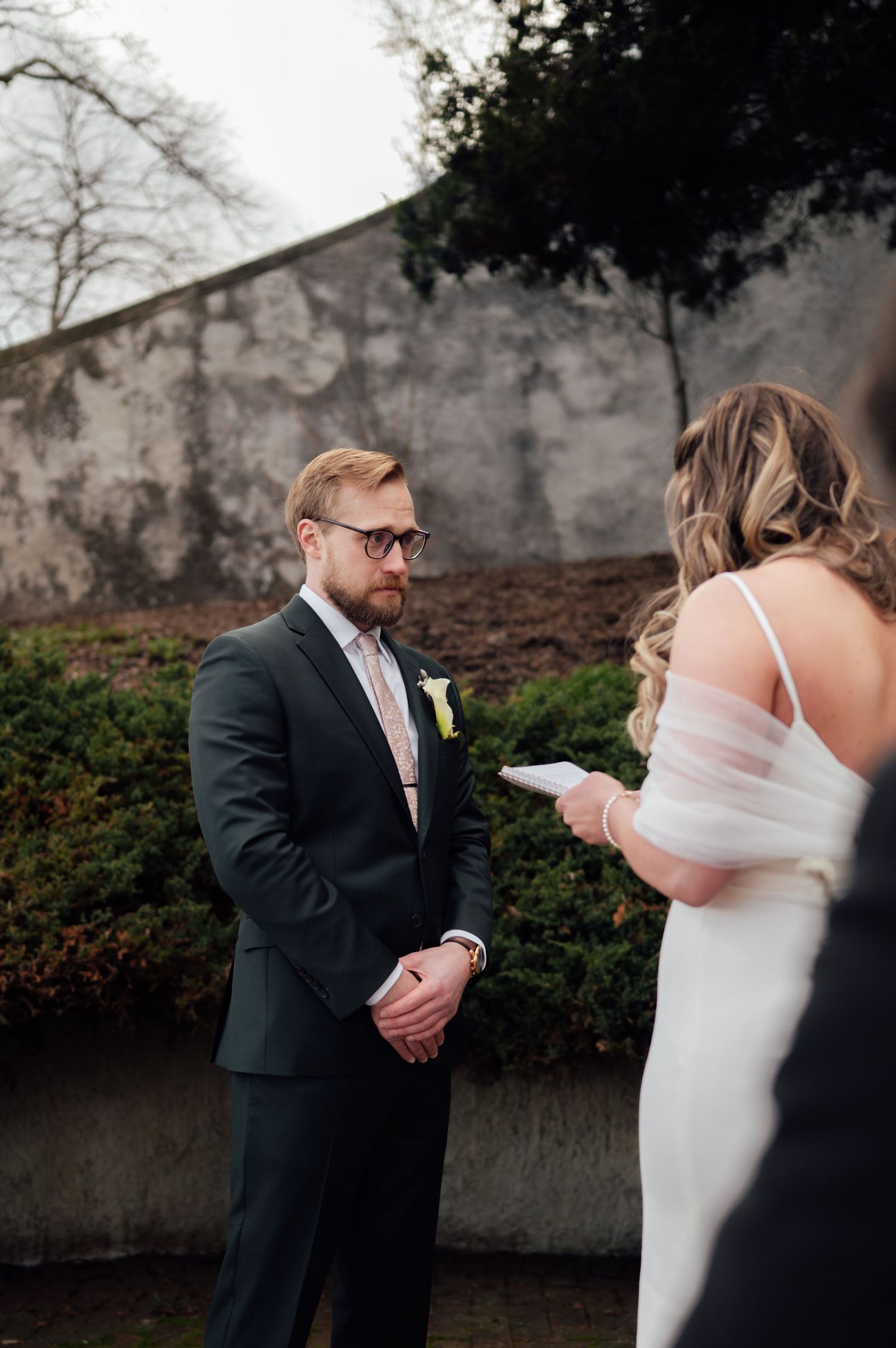 Groom in dark suit reads vows with bride in white dress during outdoor wedding ceremony.