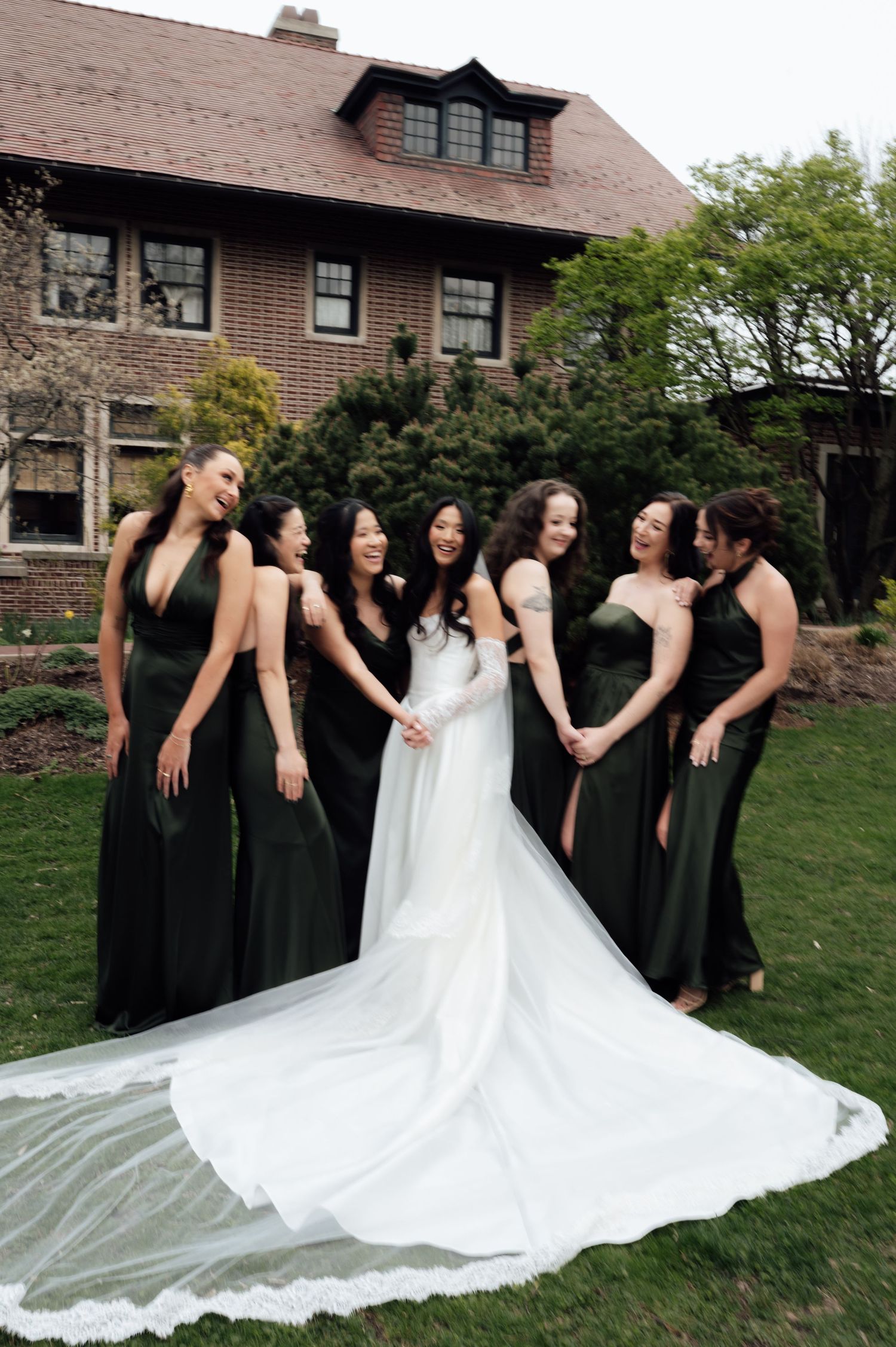 Bride in white gown with long train surrounded by bridesmaids in dark green dresses outside elegant brick mansion.