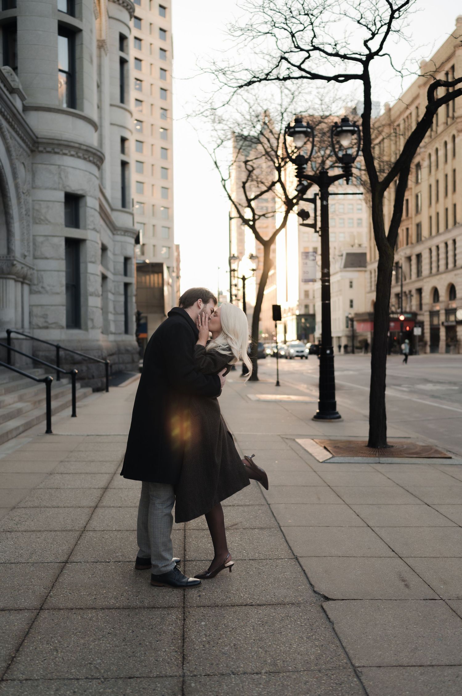 Bride and groom share a romantic kiss on a city sidewalk at sunset with urban buildings behind them.