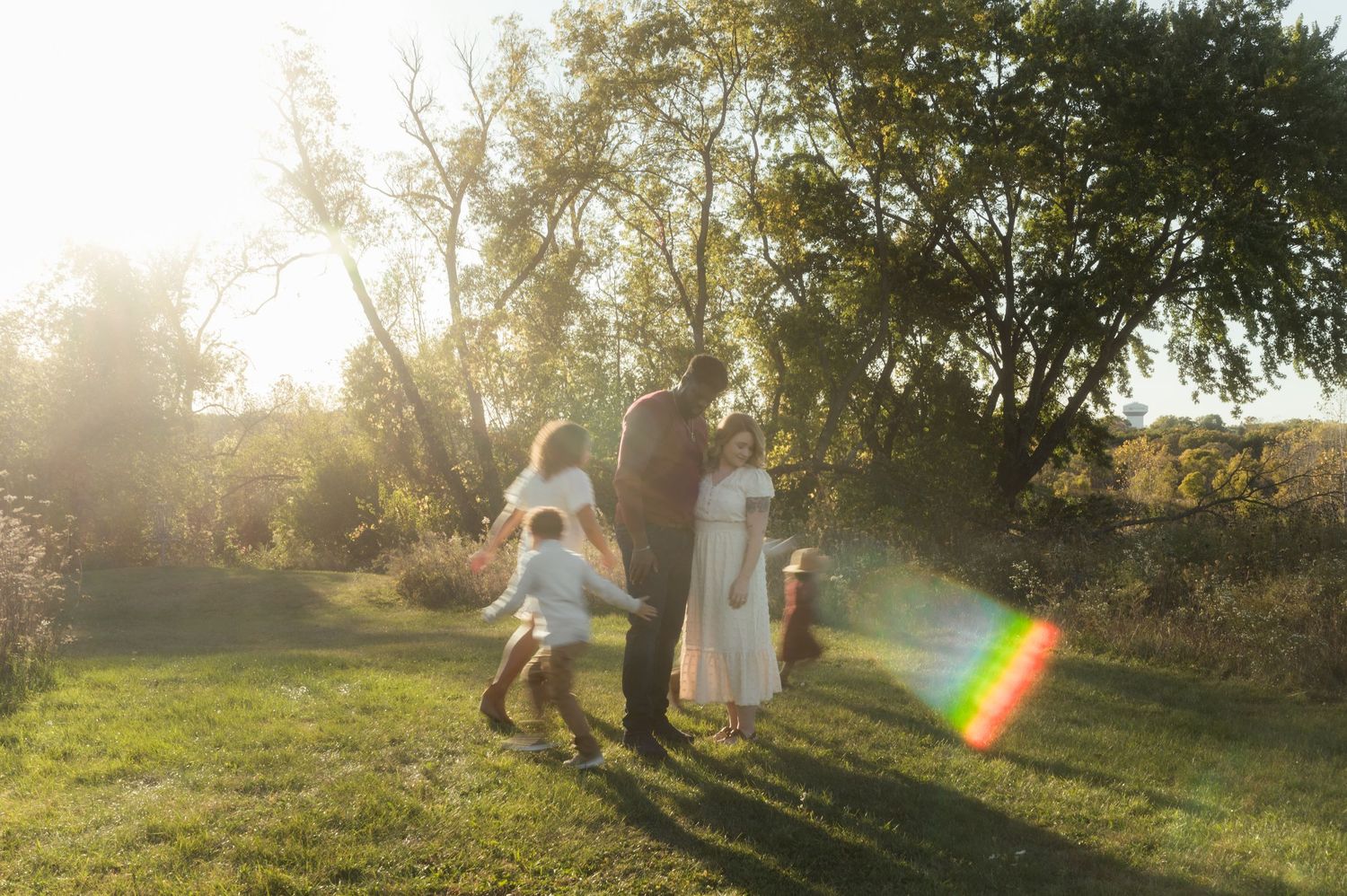 A family of four plays together in a sunlit meadow surrounded by golden autumn trees at sunset.