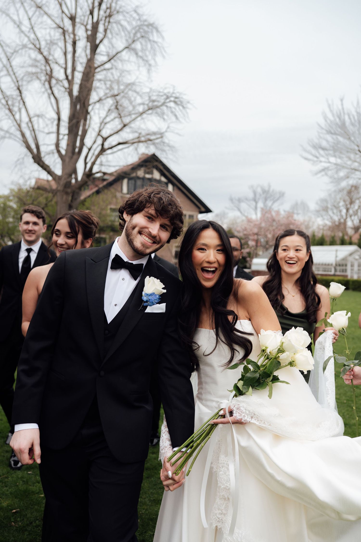 Joyful newlyweds laugh with wedding party while walking outdoors after spring wedding ceremony.