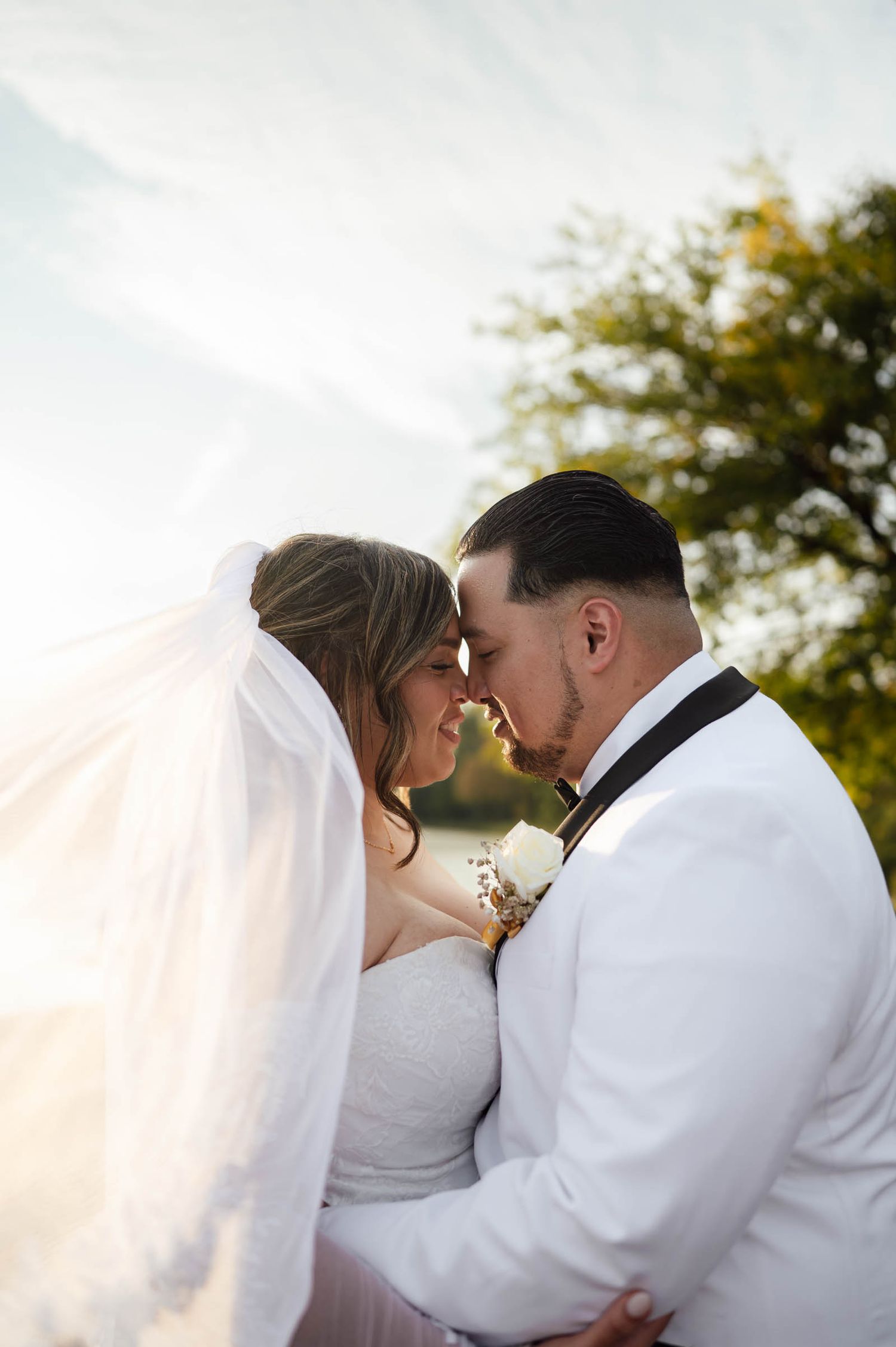 Bride and groom touching foreheads romantically with veil flowing in golden sunset light.