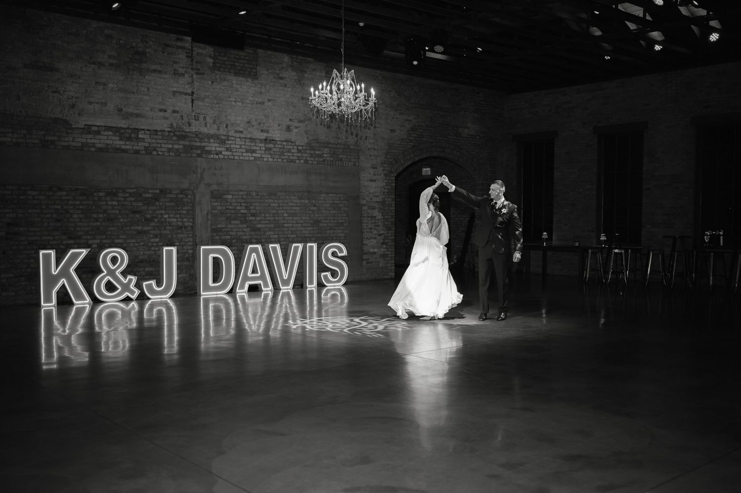 Bride and groom share first dance beside illuminated K&J Davis marquee letters in black and white photo.