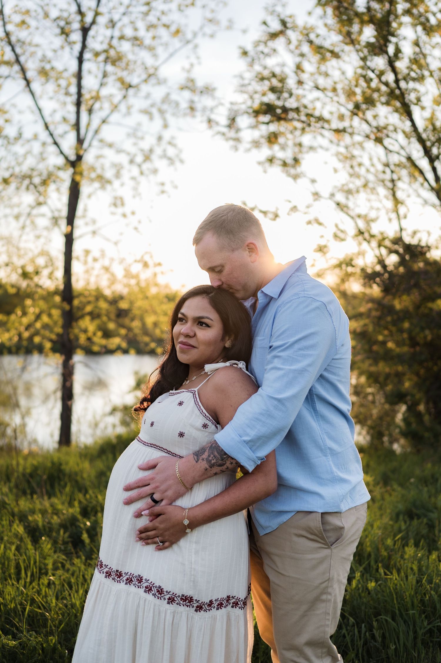 Pregnant couple embracing outdoors at golden hour by a lake, woman in white floral dress with partner holding her baby bump.