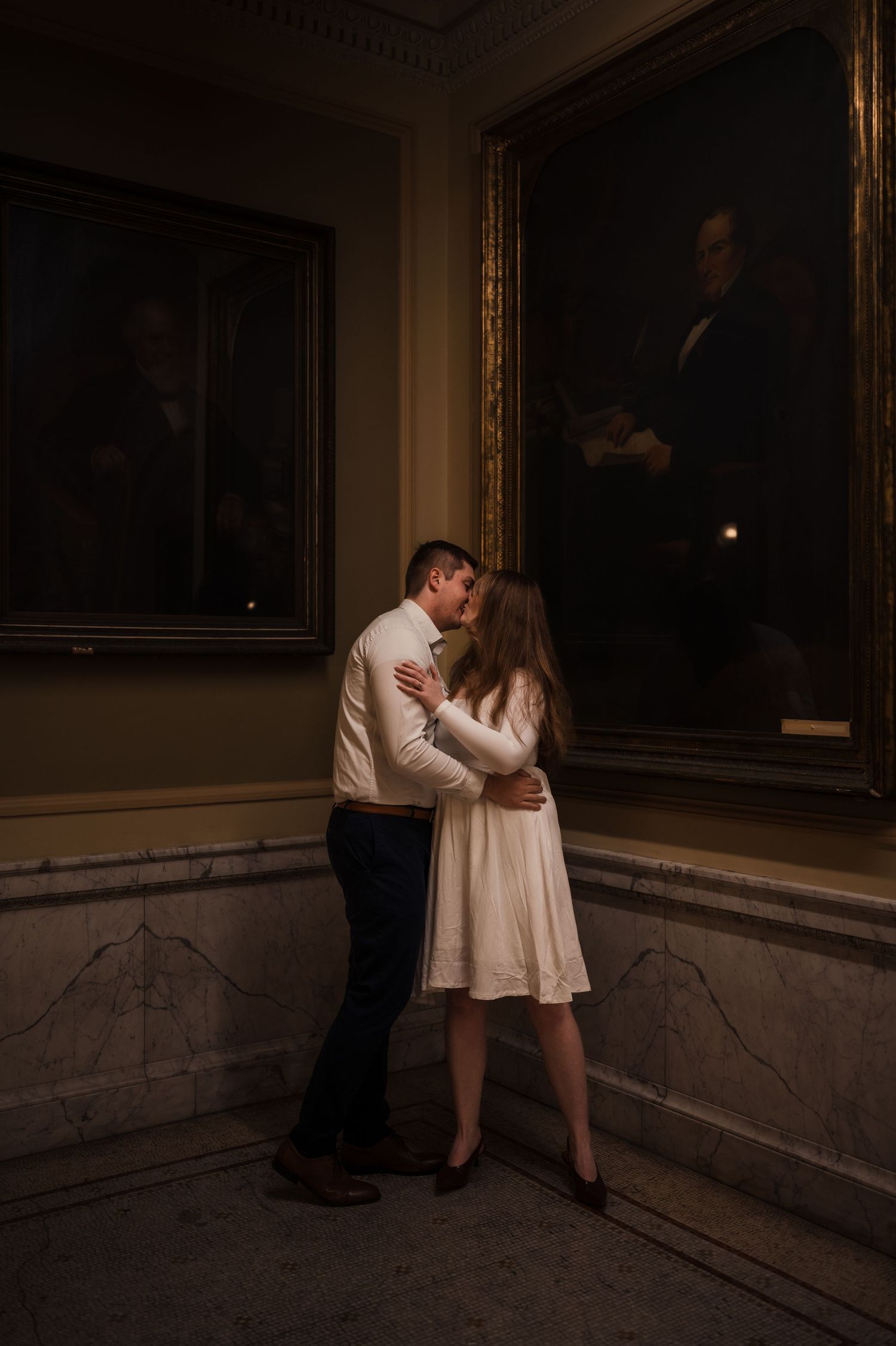 Couple sharing a romantic kiss in a dimly lit museum gallery with ornate gold-framed portrait paintings.