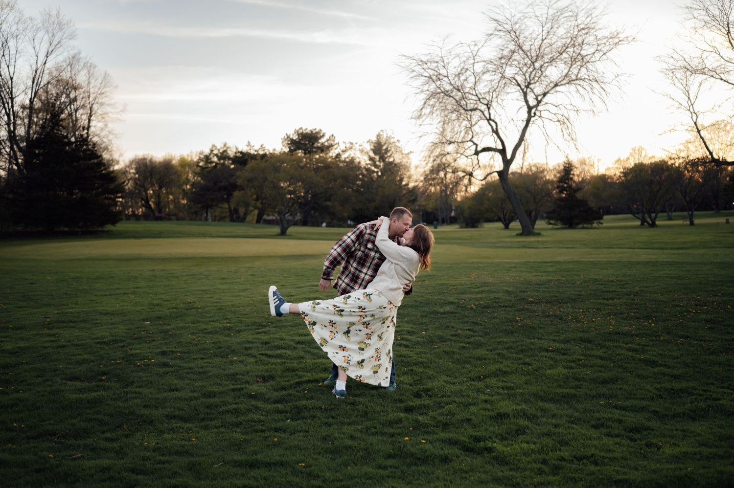 Man lifting woman in a romantic spin on a green lawn at sunset during an engagement photo session.