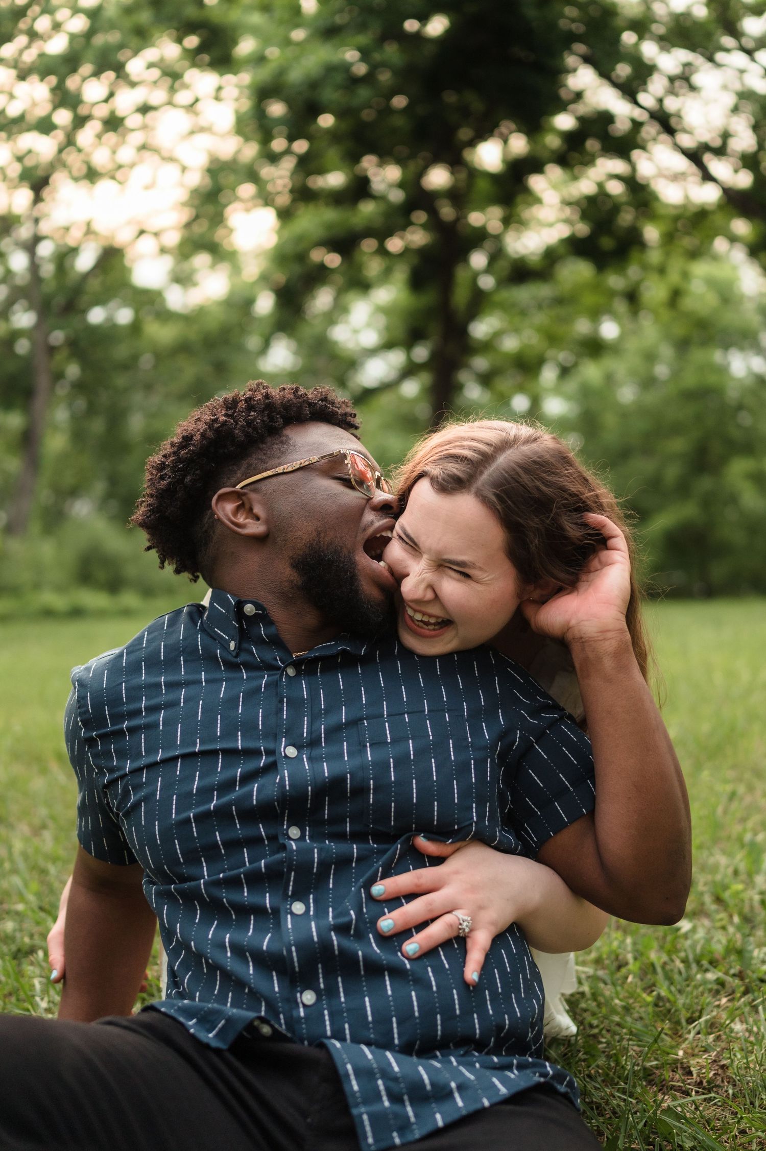 Happy interracial couple sitting on grass, man kissing womans cheek while she laughs in a park.