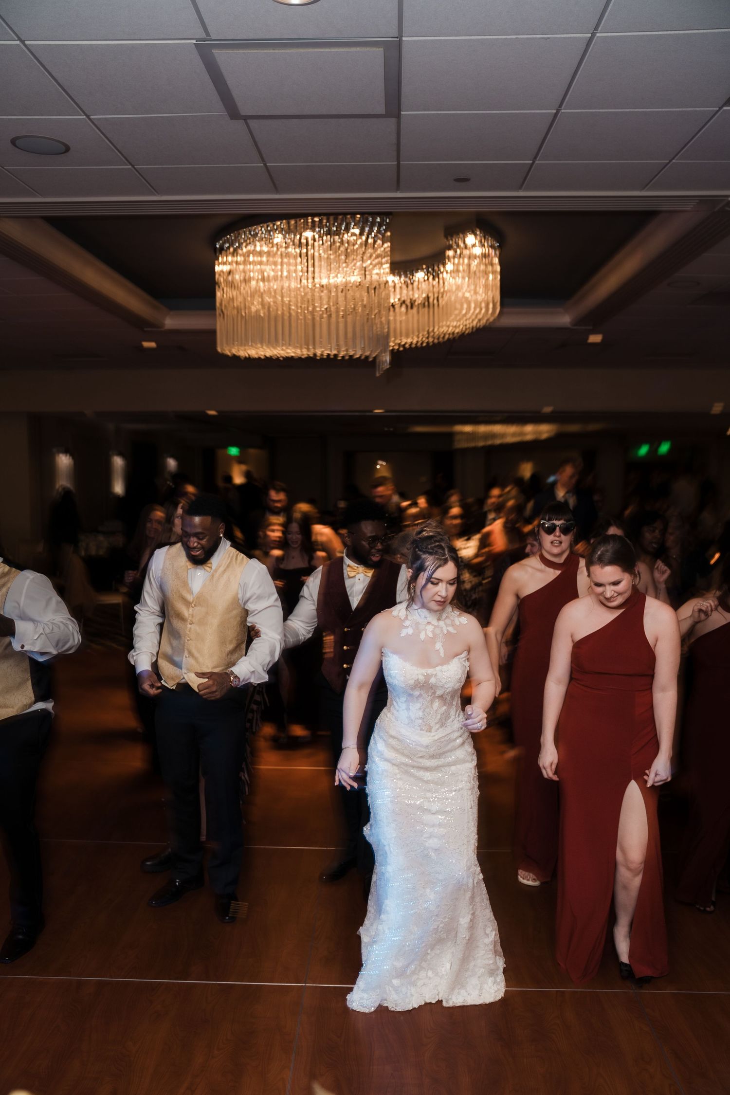 Wedding guests dance together on a dark dance floor beneath elegant crystal chandeliers at an indoor reception venue.