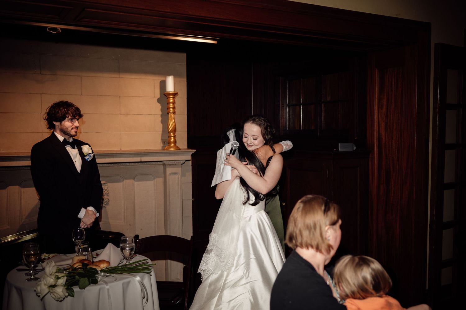 Bride in white gown hugging guest at elegant wedding reception while groom in tuxedo watches nearby.