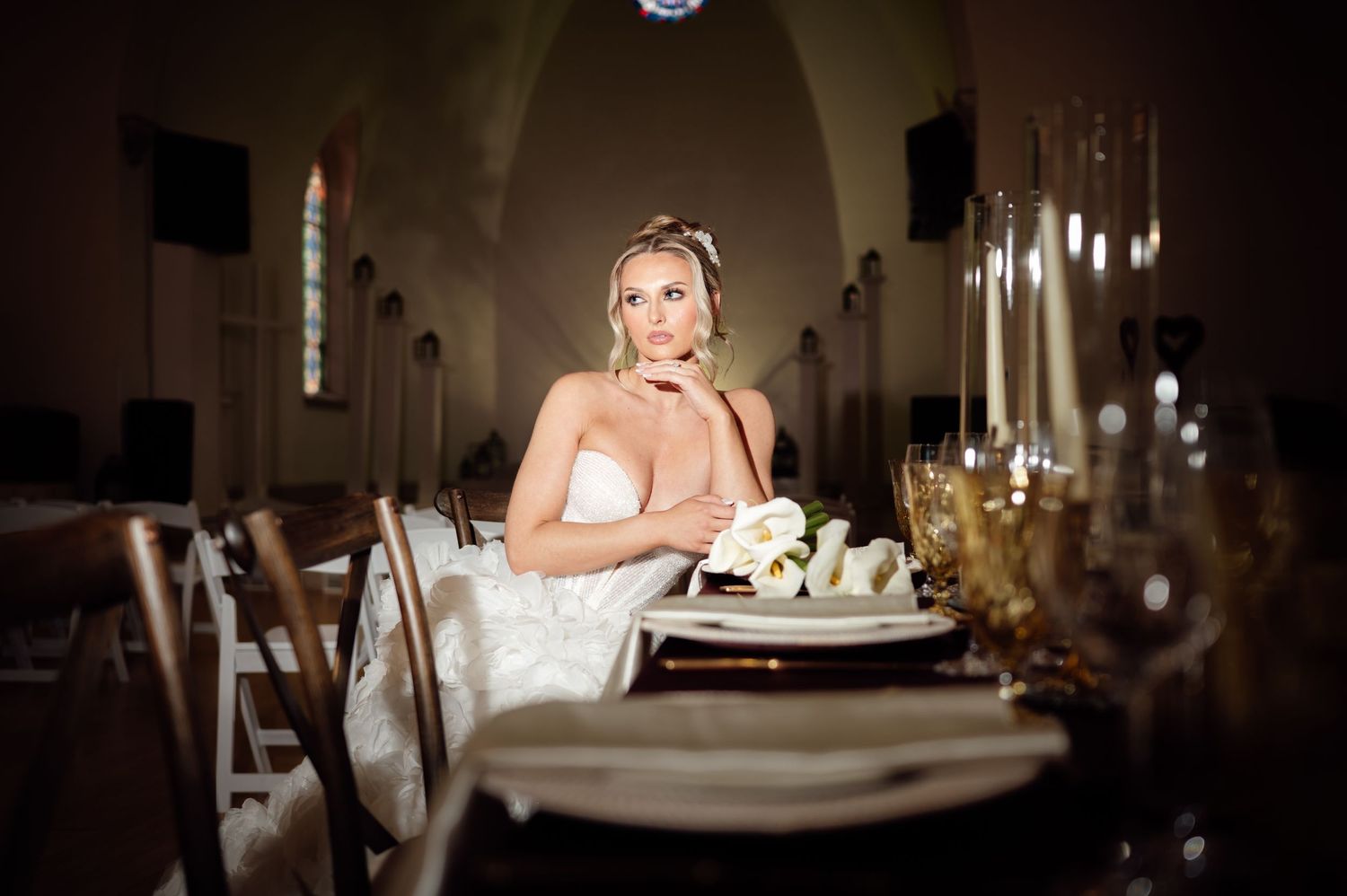 Elegant bride in strapless gown seated at a beautifully decorated wedding reception table inside a rustic church venue.
