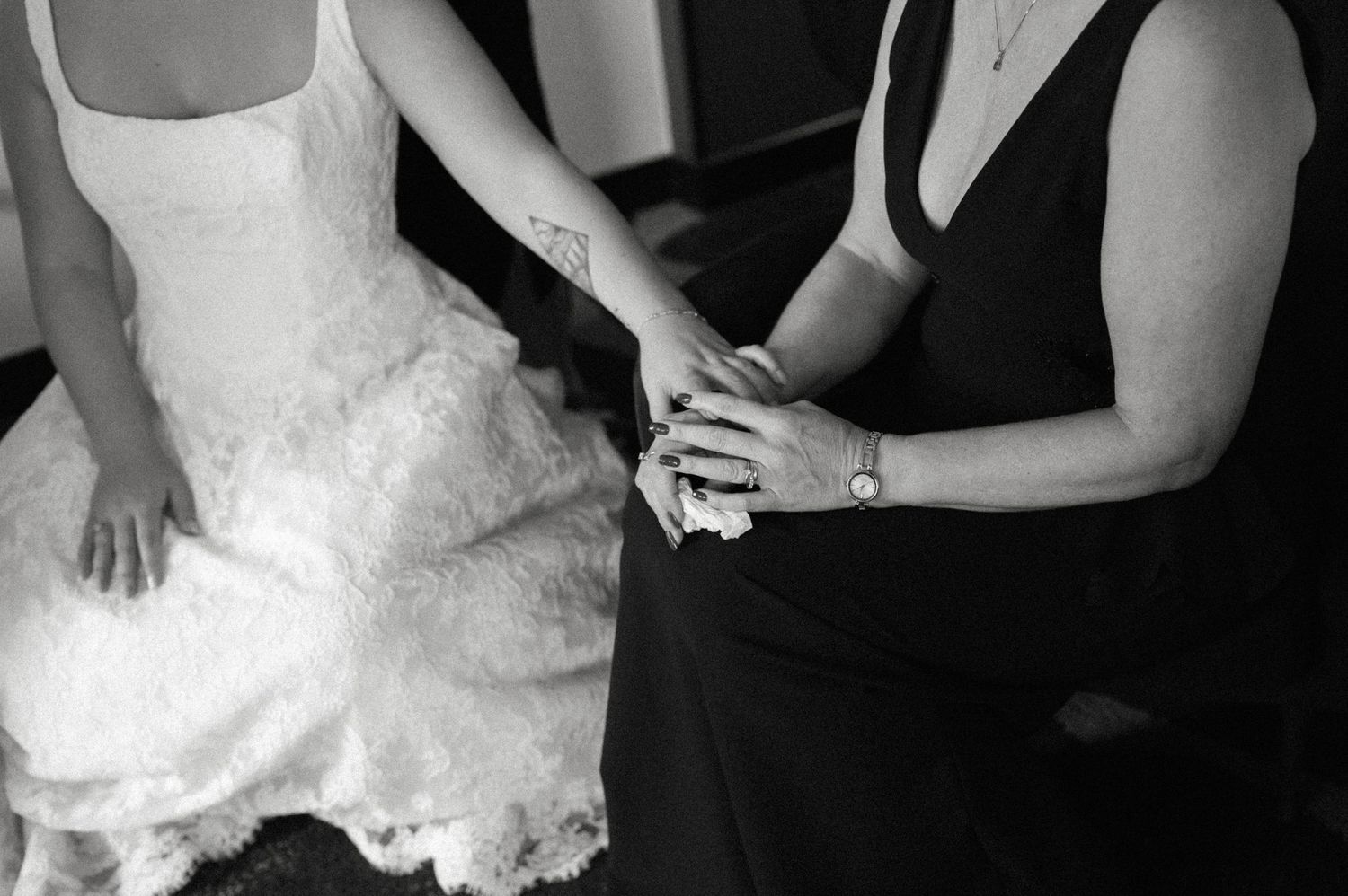 Black and white photo of bride in lace gown holding hands with woman in black dress at wedding.