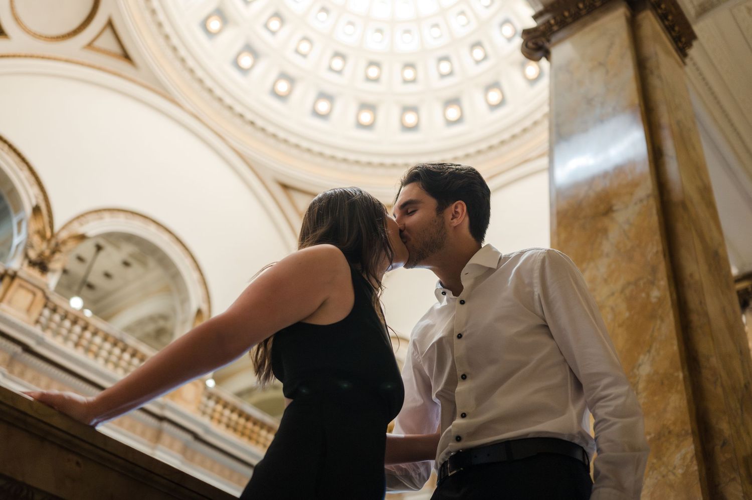 Couple sharing a romantic kiss on grand staircase inside an elegant domed marble building.