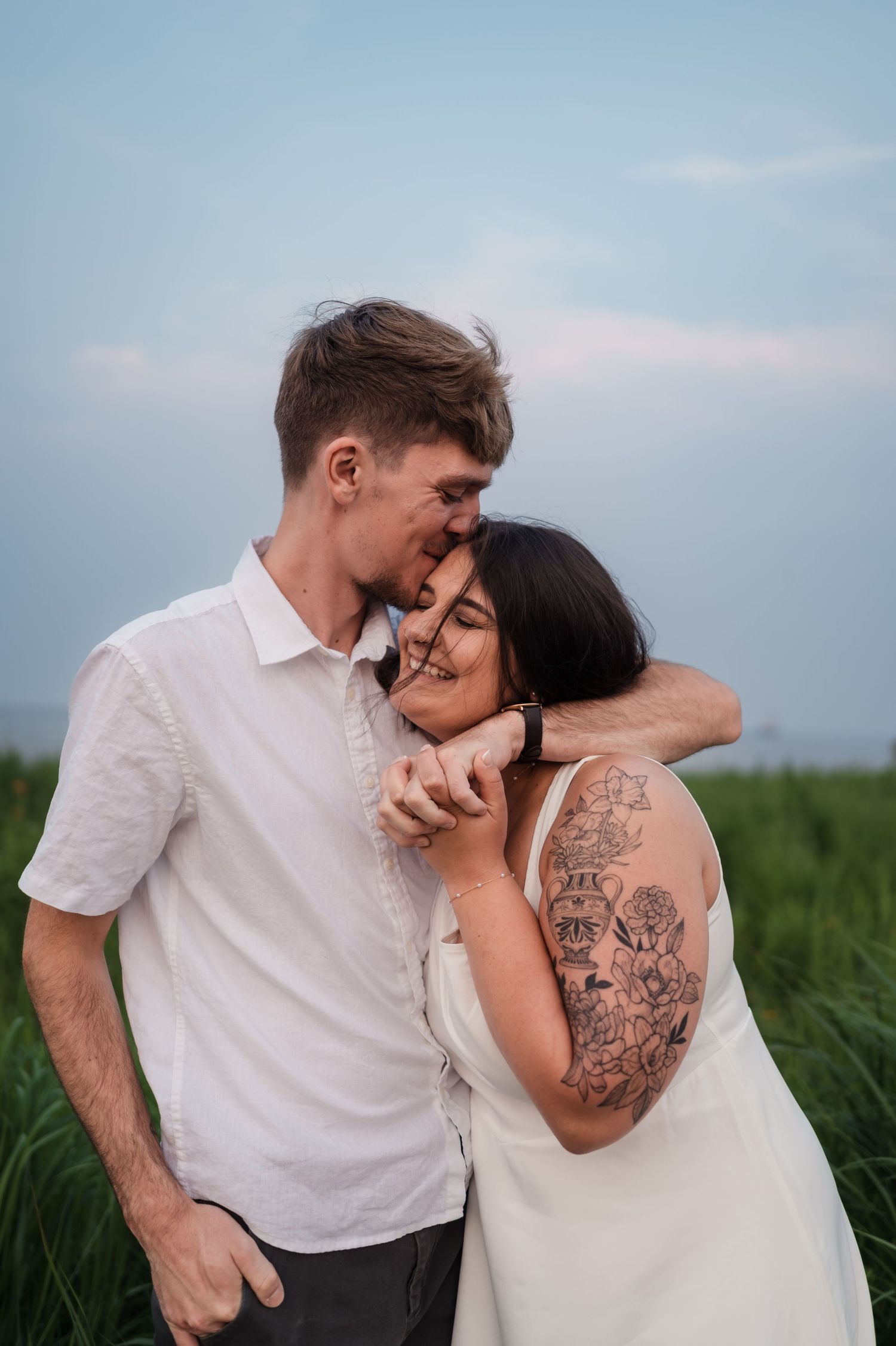 Man kissing woman on forehead in a green field, she smiles while leaning into his embrace.