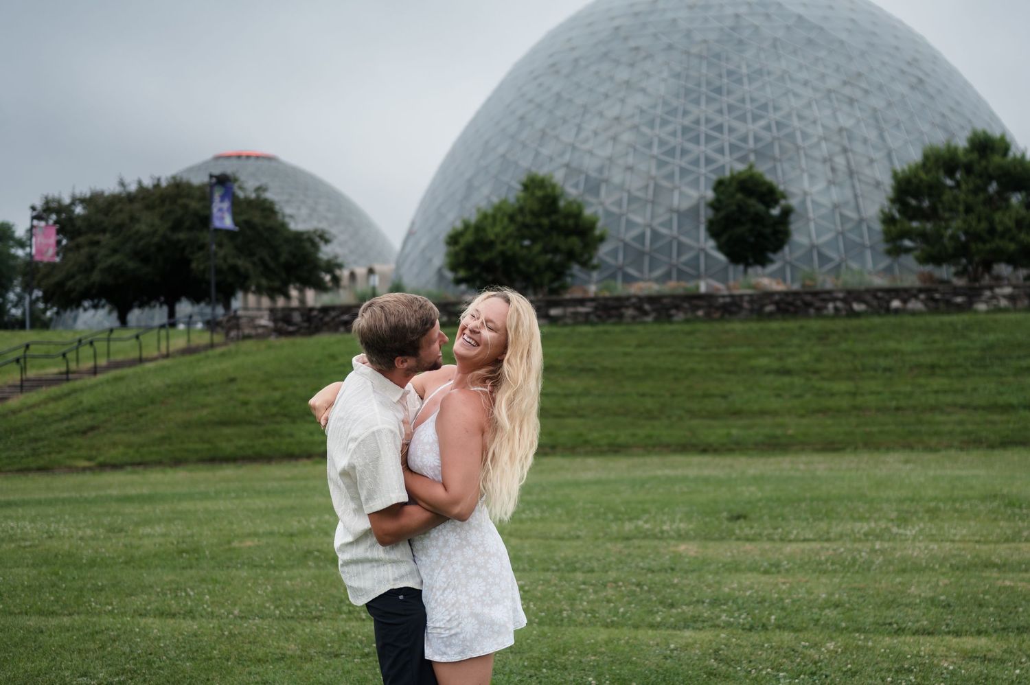 Happy couple embracing and laughing on a green lawn with geodesic dome structures in background.