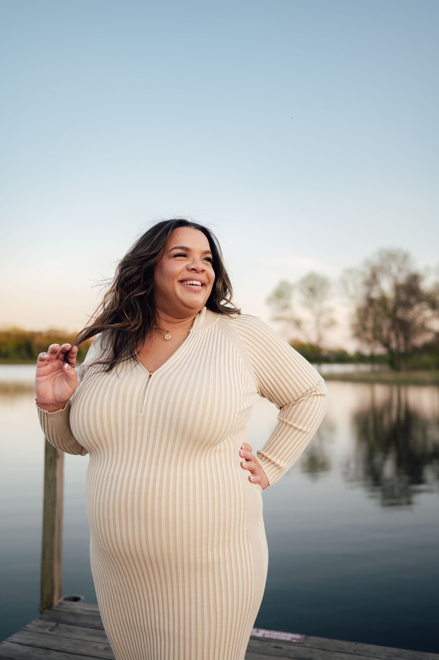 Smiling pregnant woman in a beige ribbed dress posing on a dock at sunset by a lake.