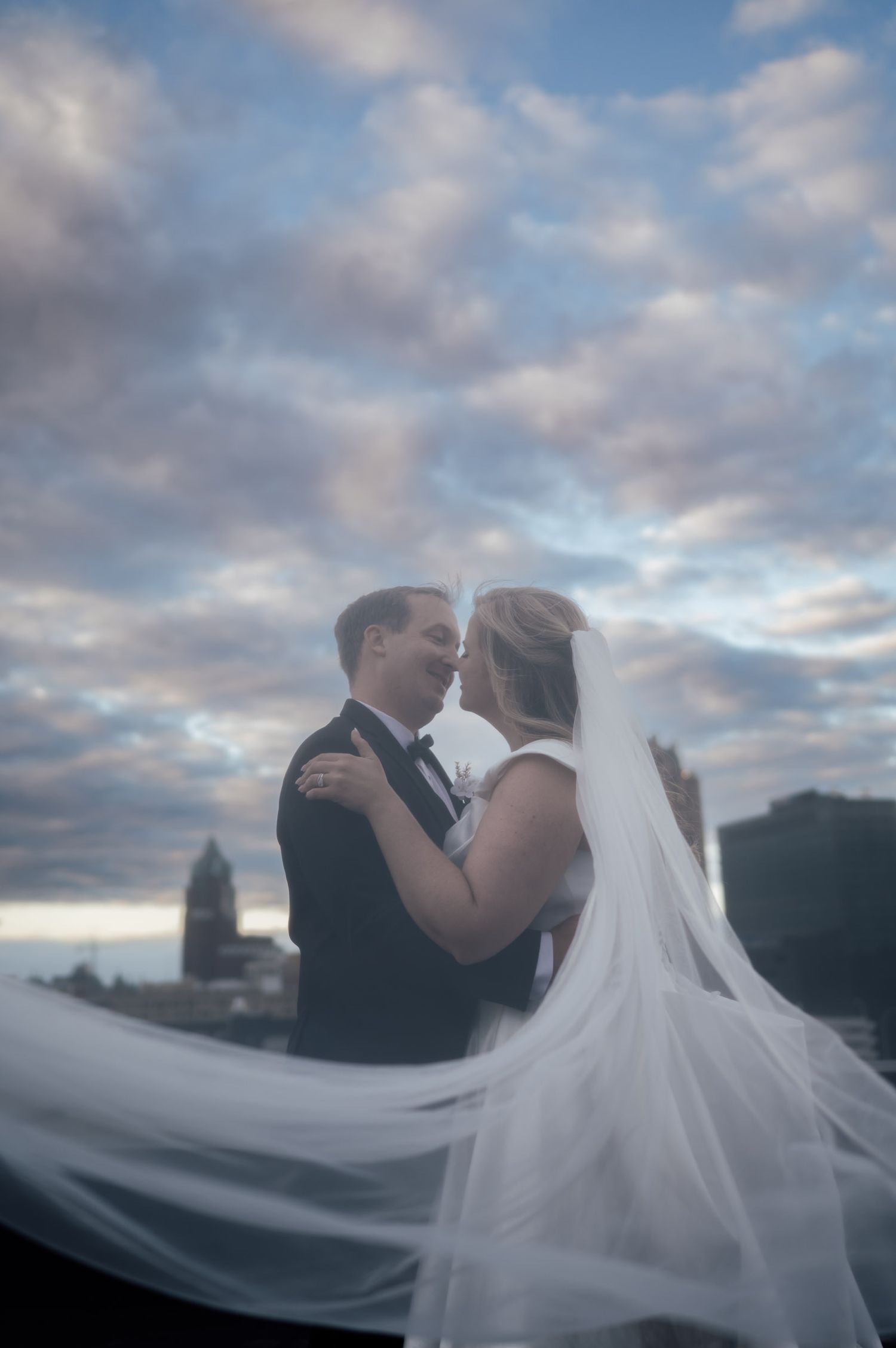 Romantic bride and groom nearly kissing with veil flowing in the wind against a dramatic cloudy sunset sky.