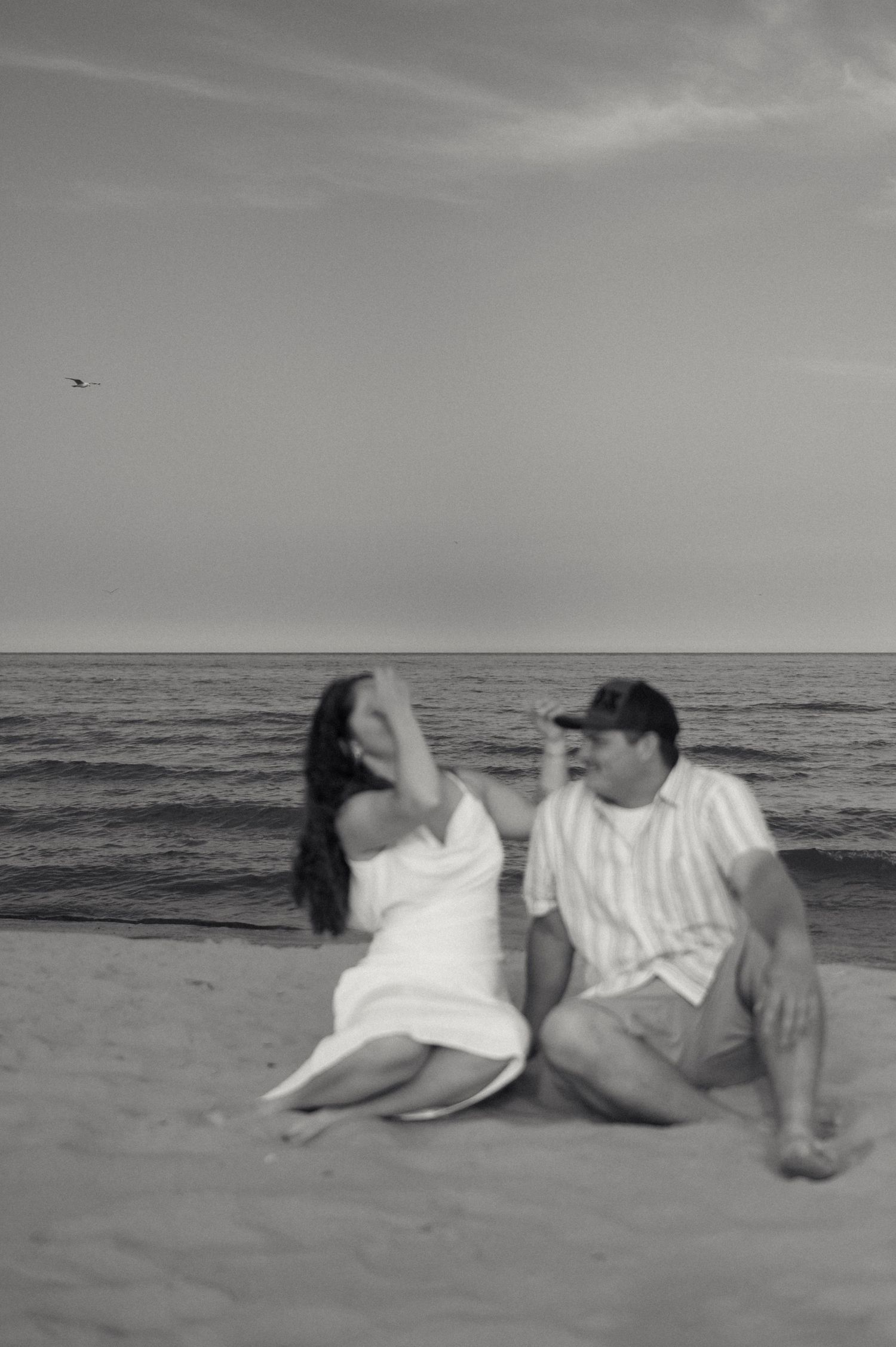 A couple sits together on a sandy beach at sunset in this black and white photograph.