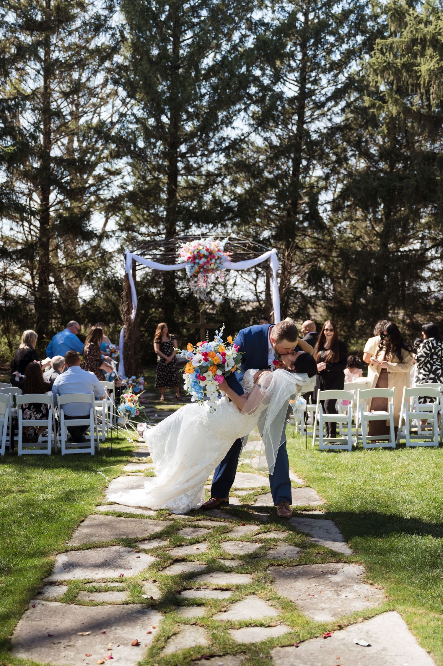An outdoor garden wedding ceremony with a floral arch, white chairs and stone pathway surrounded by tall evergreen trees.