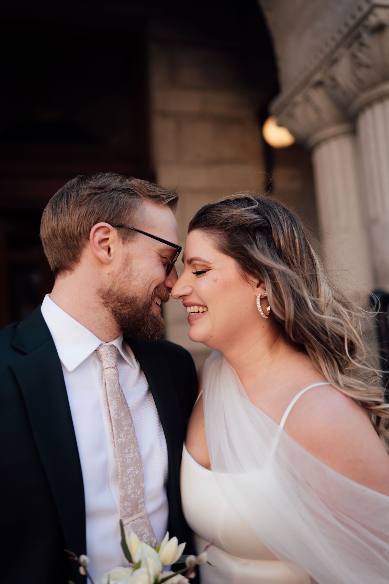 A smiling bride and groom touch foreheads tenderly outside a grand building on their wedding day.
