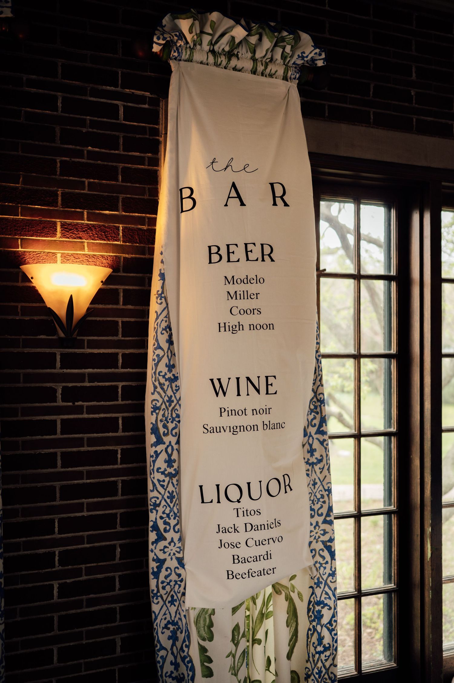 Elegant bar menu banner listing beer, wine, and liquor options hanging near a window in a dimly lit brick venue.