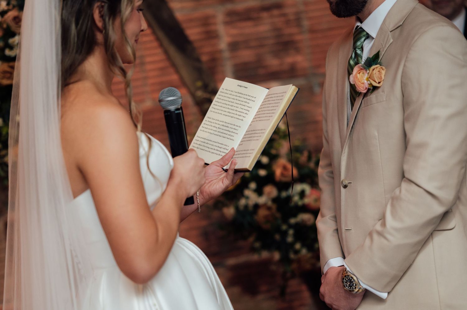 Bride reading vows from a book during wedding ceremony, wearing white gown with veil, groom in tan suit beside her.