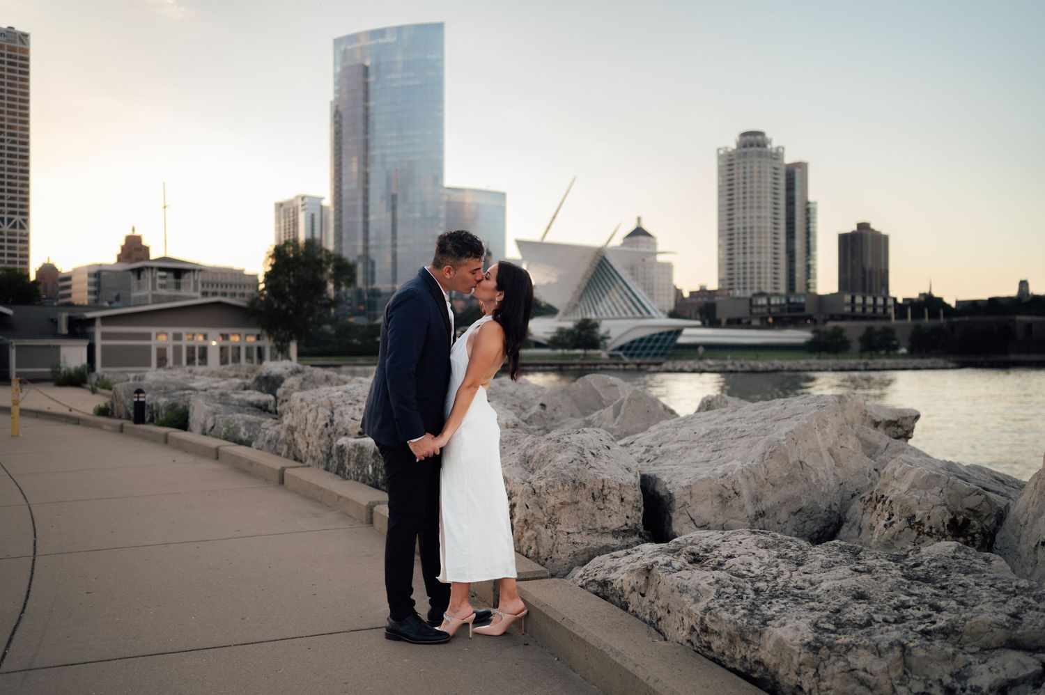 Couple sharing a romantic kiss on a lakeside promenade with Milwaukee skyline at sunset.