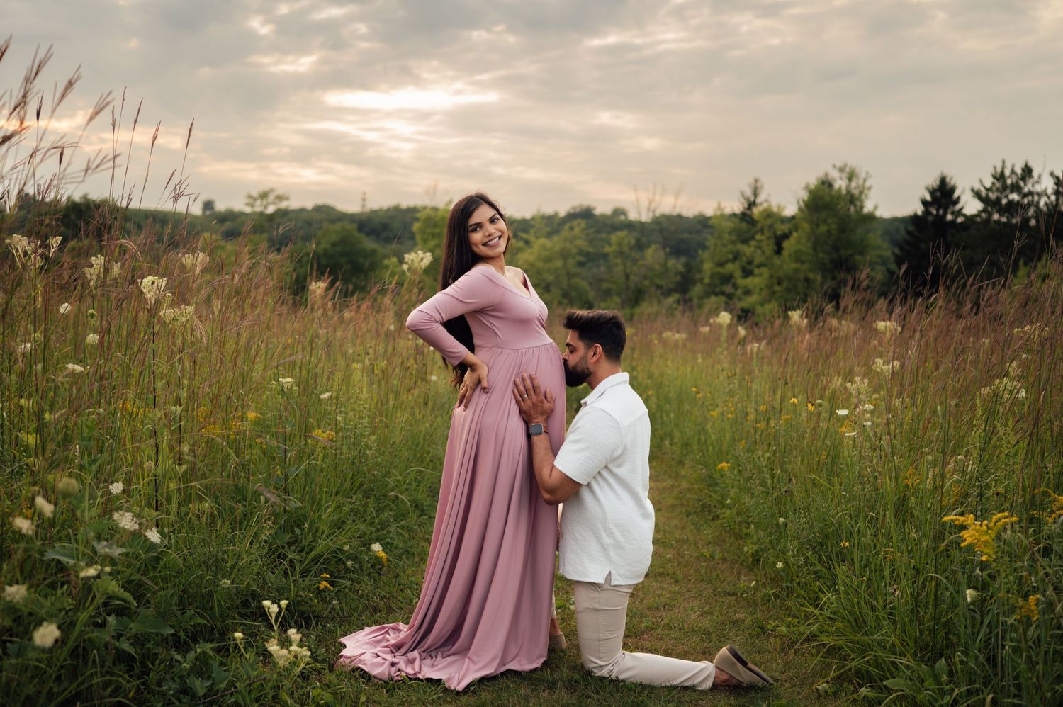 A man kneels before a pregnant woman in a pink maternity gown in a lush green meadow at sunset.