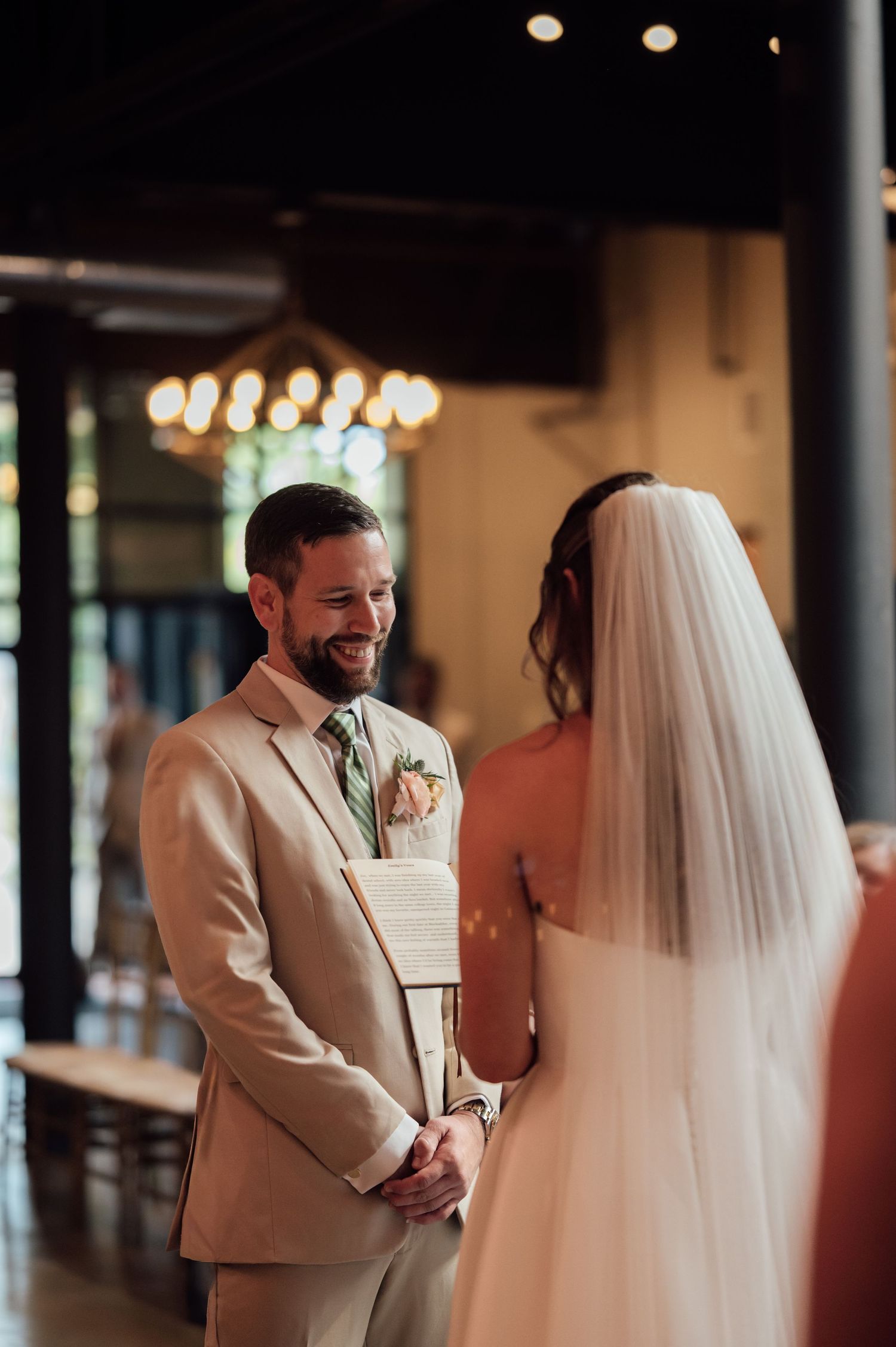 Groom in tan suit smiling at bride in white veil during indoor wedding ceremony with warm chandelier lighting.