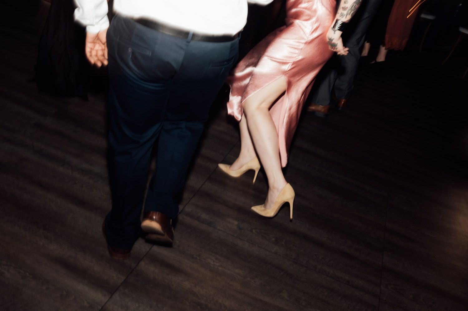 Couple dancing at wedding reception, woman in pink satin dress and nude heels on dark floor.