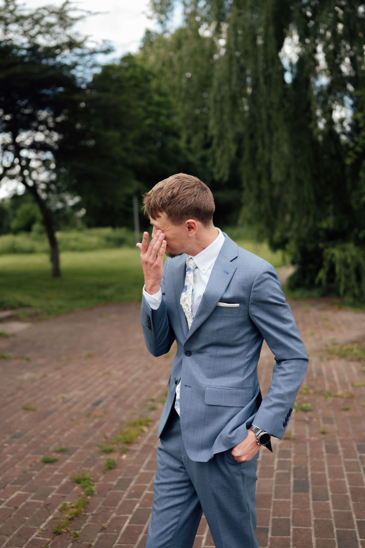 Emotional groom in blue suit covering face with hand on brick path surrounded by lush green trees.