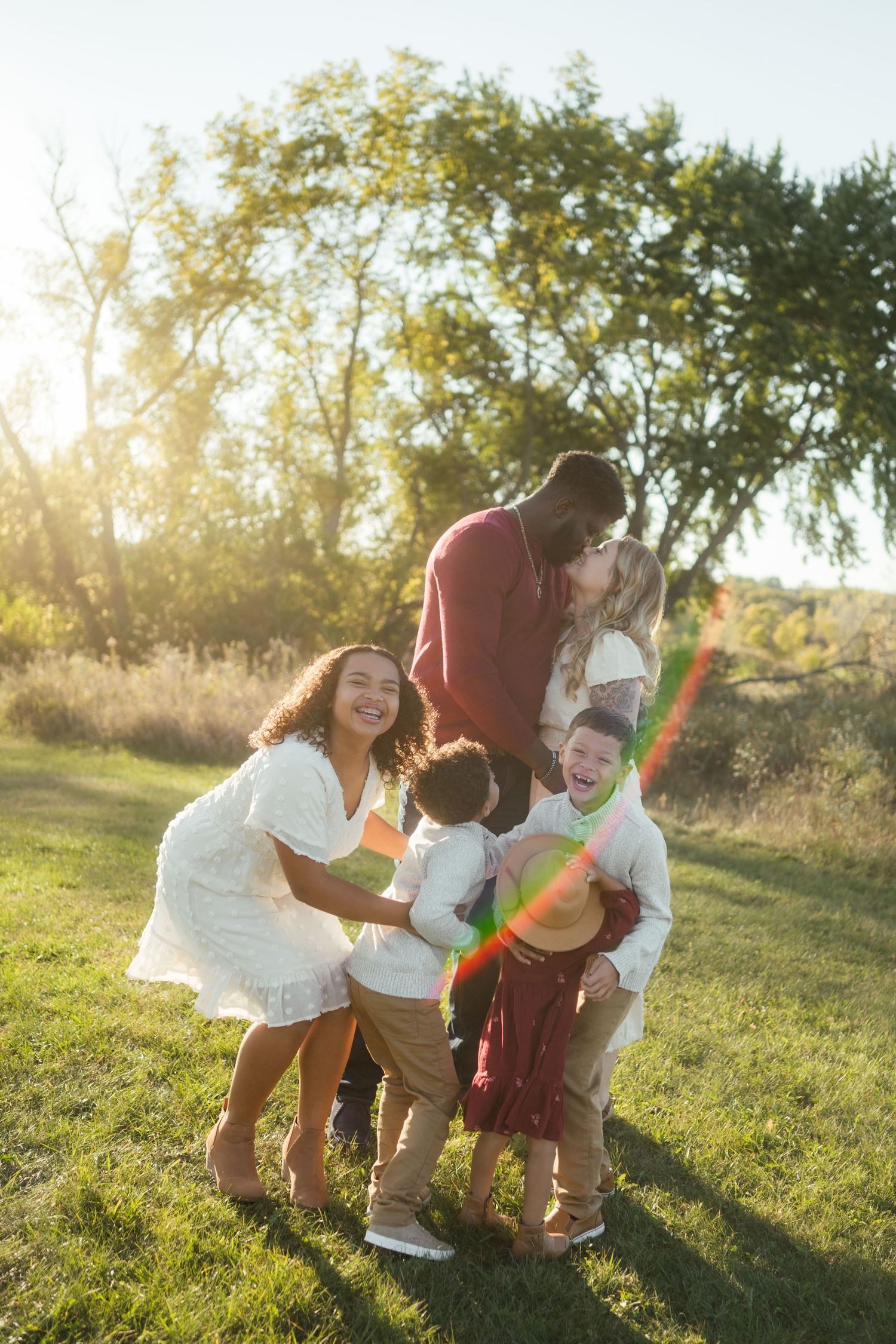 Joyful blended family laughing and playing together on green grass with bright sun flare.