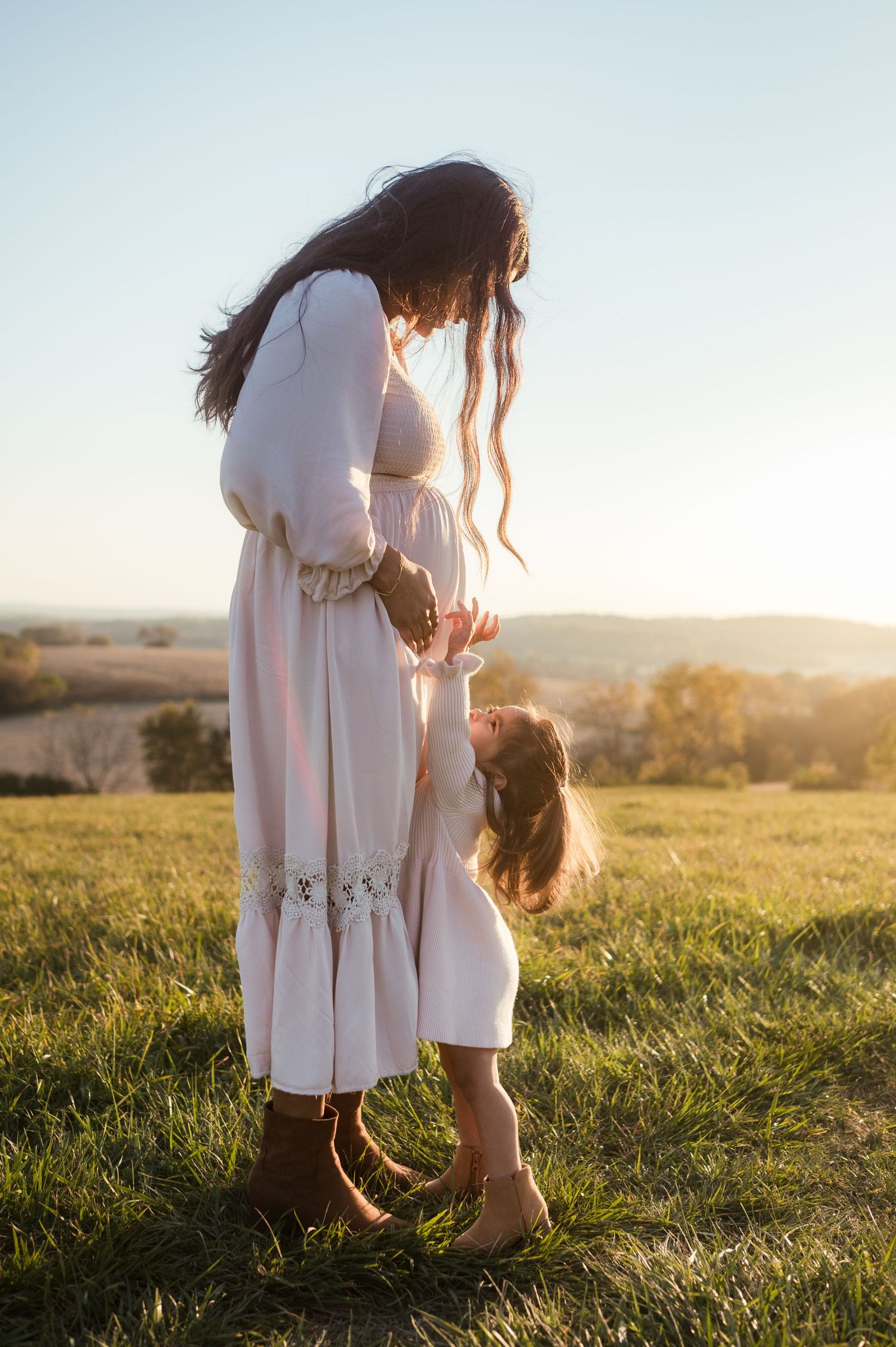 Pregnant mother in pink dress holding daughters hands at golden hour on grassy hilltop.