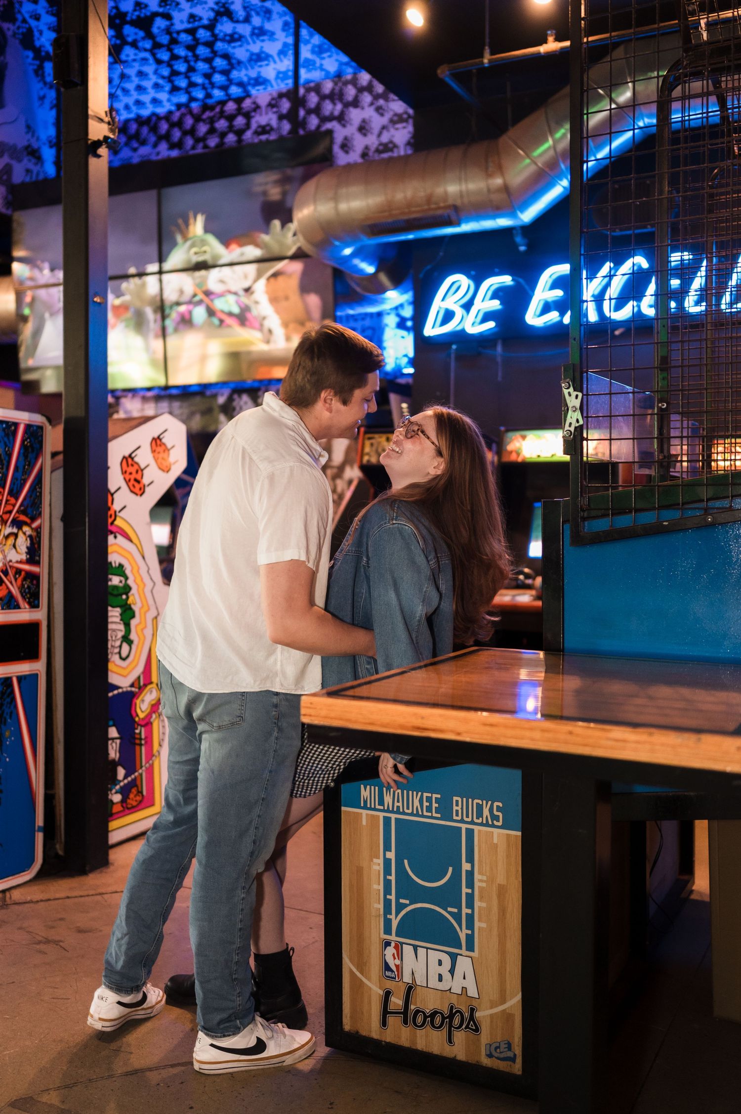 Couple sharing a sweet kiss while leaning against an NBA Hoops game inside a colorful arcade bar.