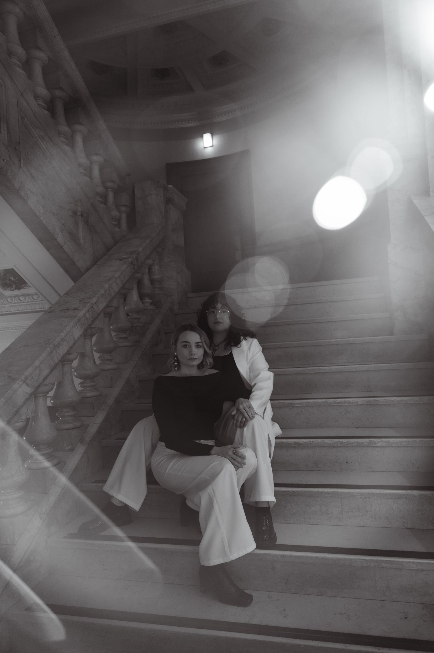 Black and white photo of two women sitting closely together on grand marble stairs.