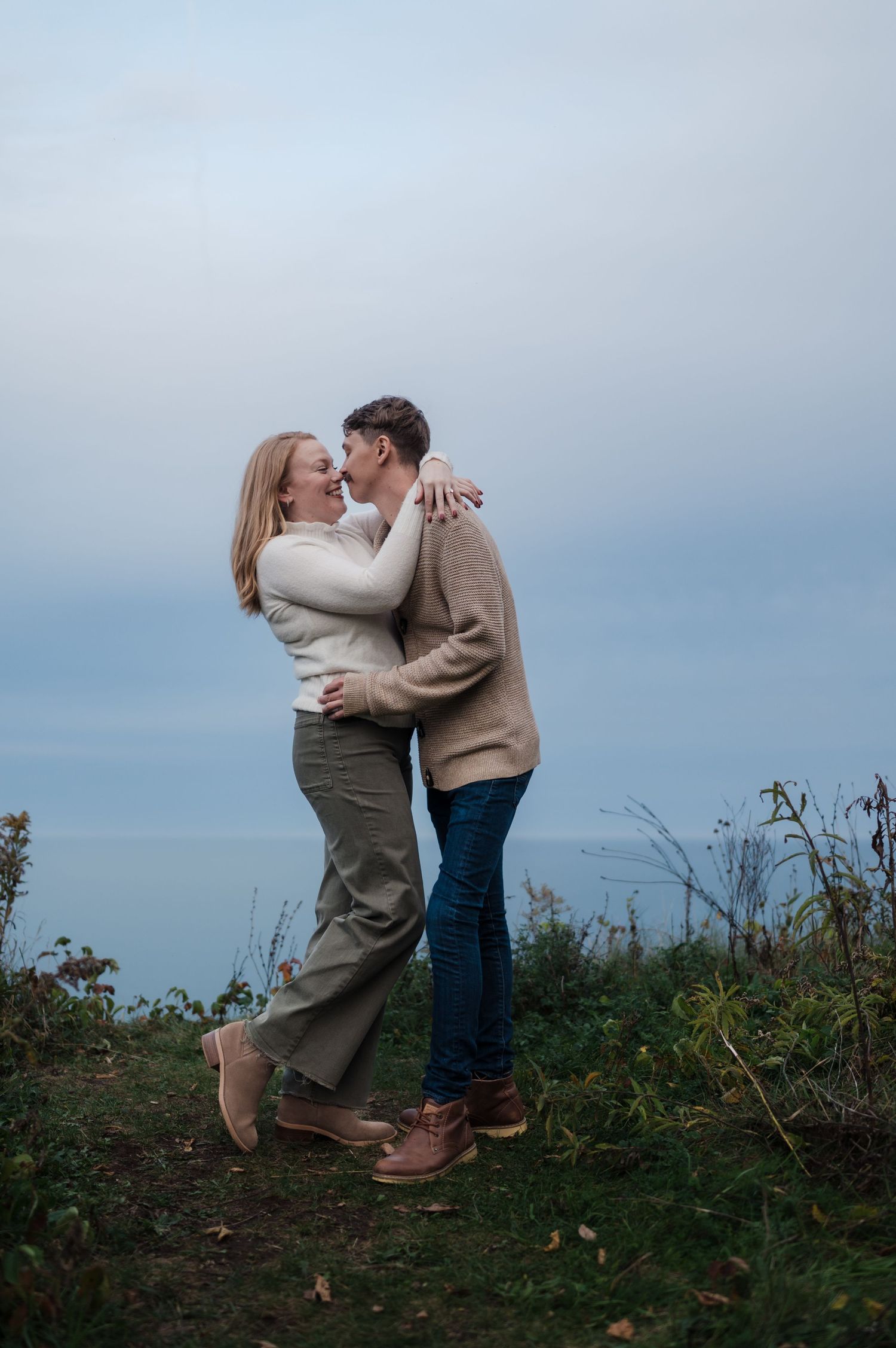 Joyful couple leaning in for a kiss on a scenic overlook with overcast skies in the background.