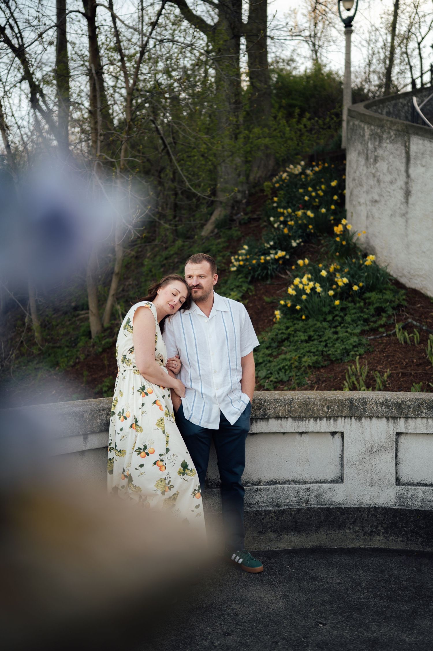 Couple embracing by a stone wall with spring daffodils blooming in the background on a soft day.