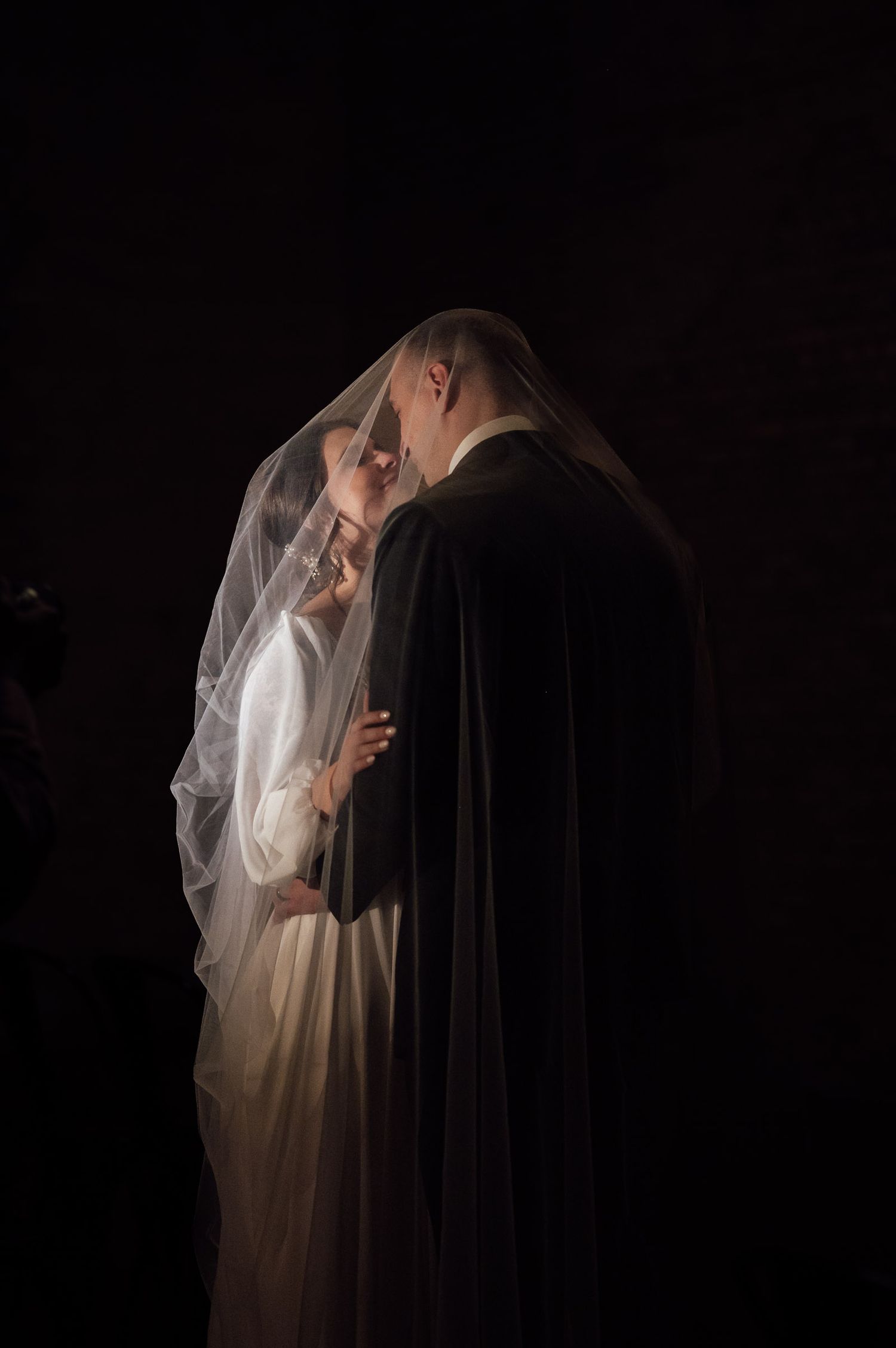 Bride and groom share an intimate kiss under a sheer wedding veil against a dark background.