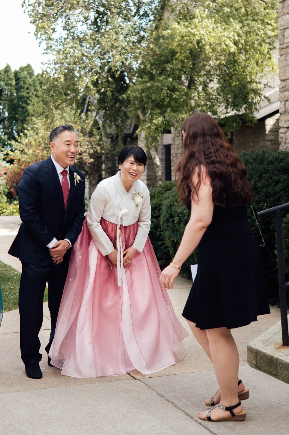 Woman in traditional Korean pink hanbok greeted by guests outside wedding venue.