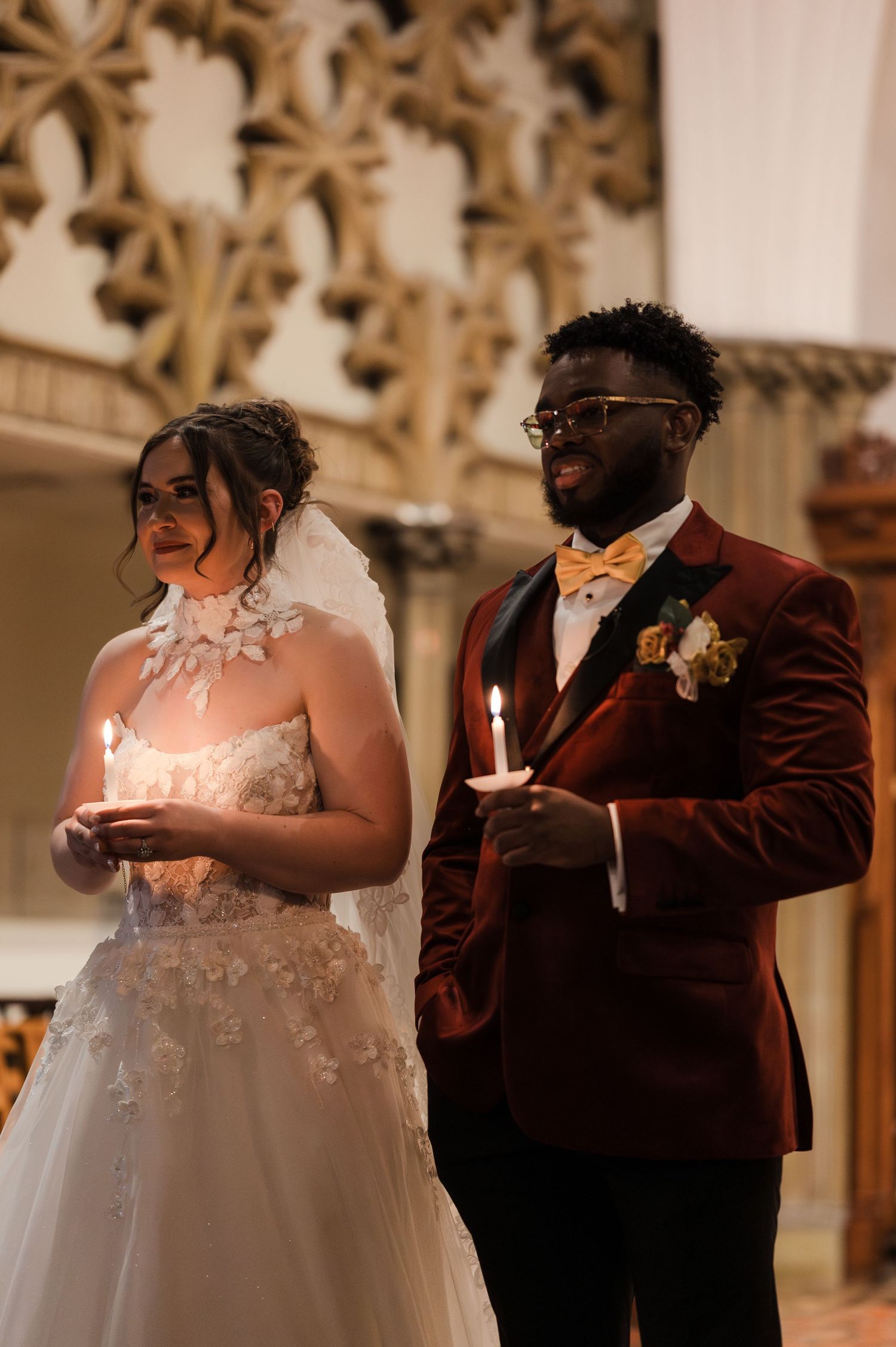 Bride in lace gown and groom in burgundy velvet suit hold candles during wedding ceremony in ornate church.