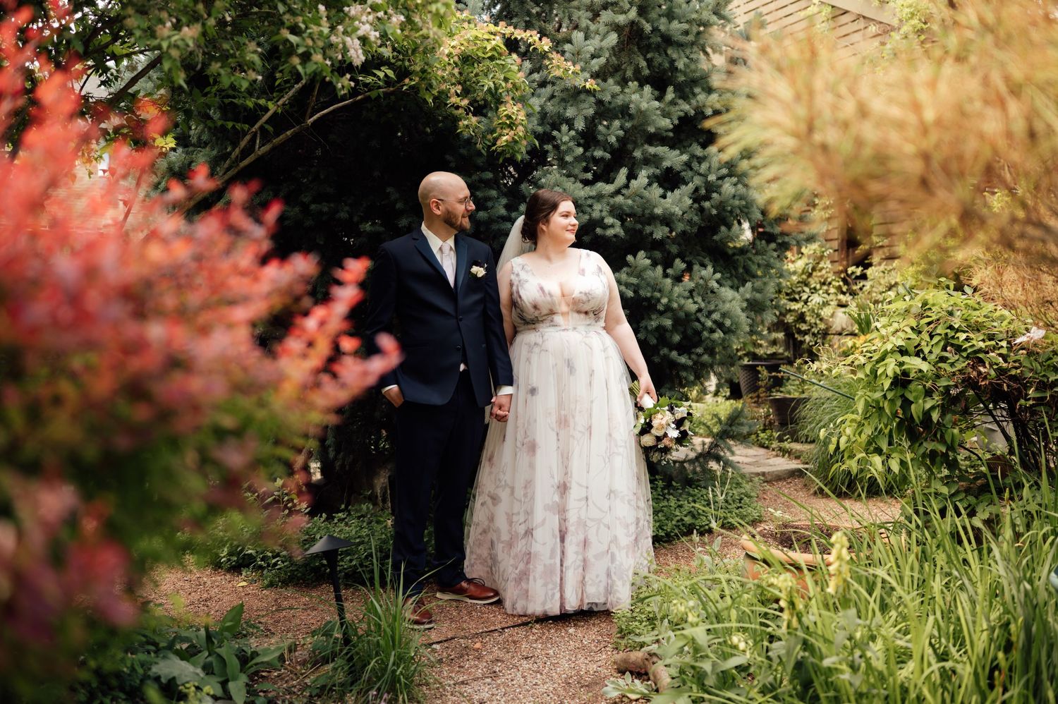 Bride in floral gown and groom in navy suit walk hand in hand through a lush garden path surrounded by colorful foliage.