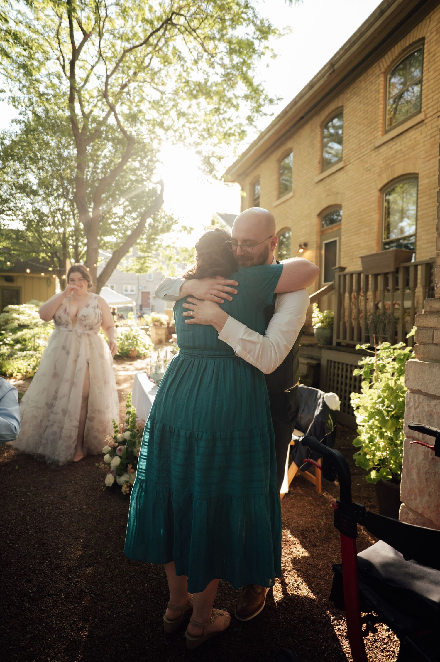 Bald groom embracing guest in teal dress at outdoor wedding with bride visible in background.
