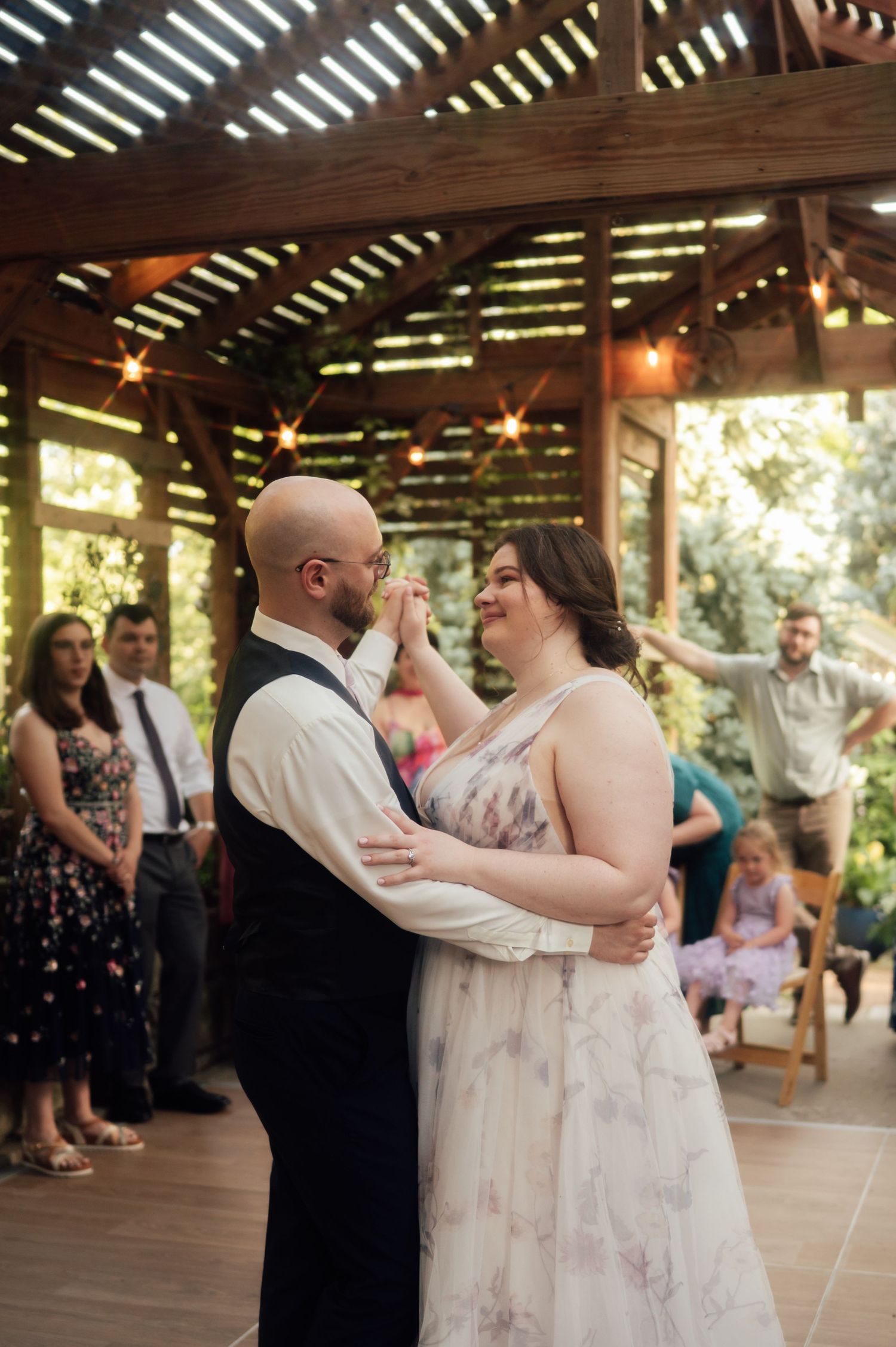 Couple shares first dance at outdoor wedding reception under pergola with string lights and guests watching.