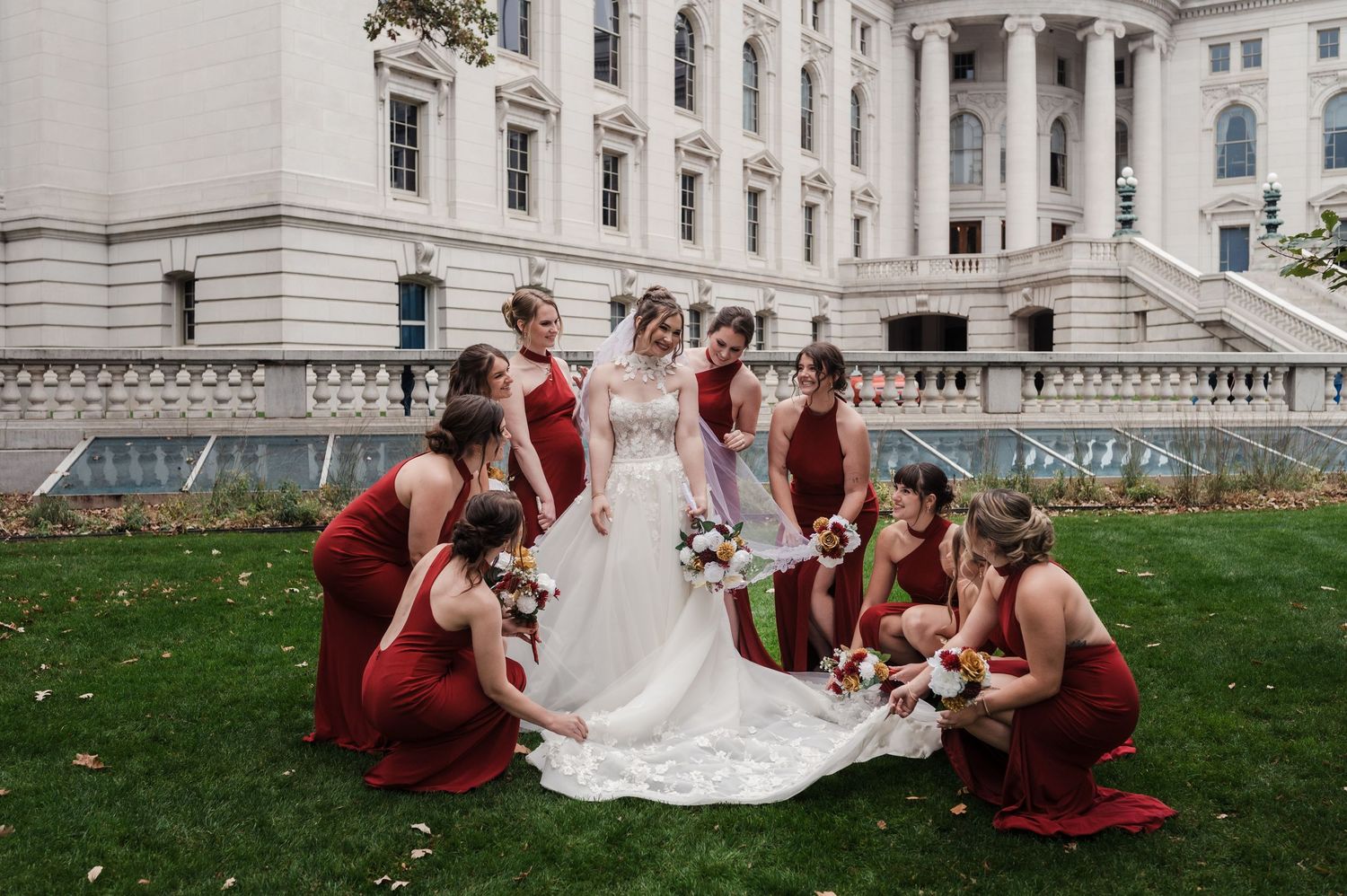A wedding party in burgundy dresses and white gown poses on the steps of a grand stone building.