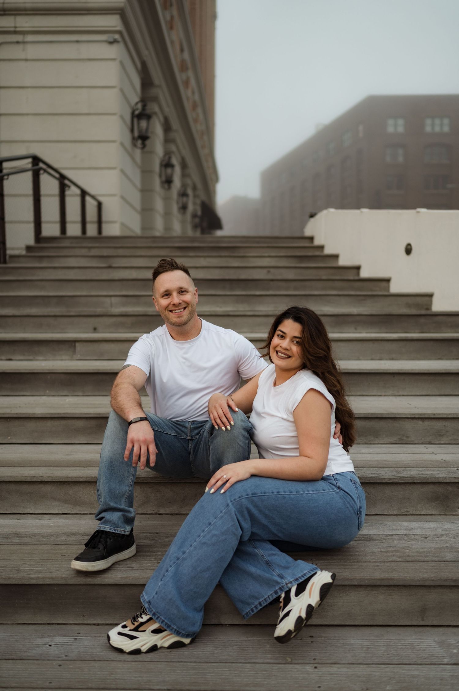 A couple wearing white t-shirts and jeans sit together on concrete steps outside a building on a foggy day.