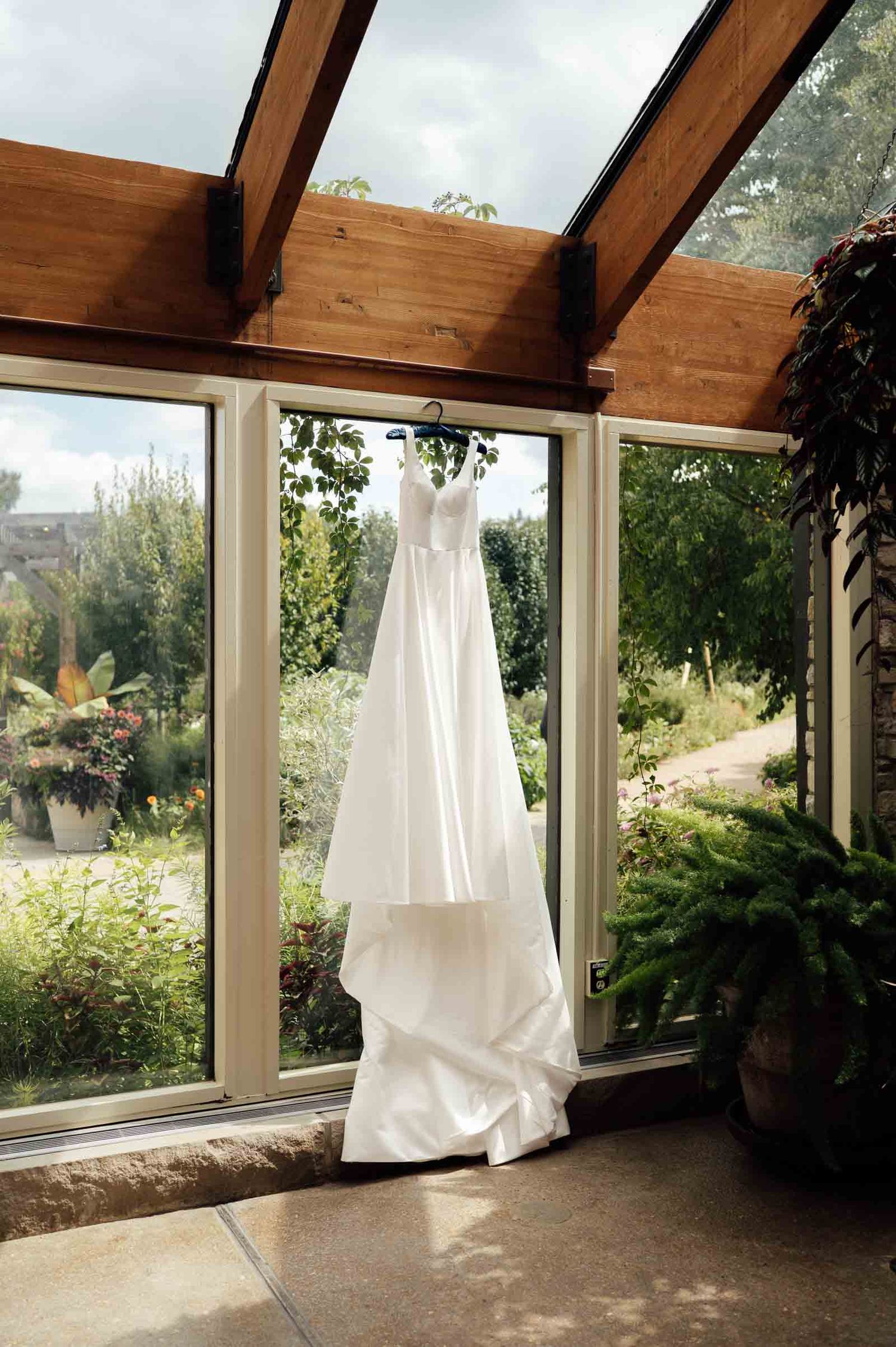 White wedding dress hanging in sunlit greenhouse window surrounded by lush greenery.