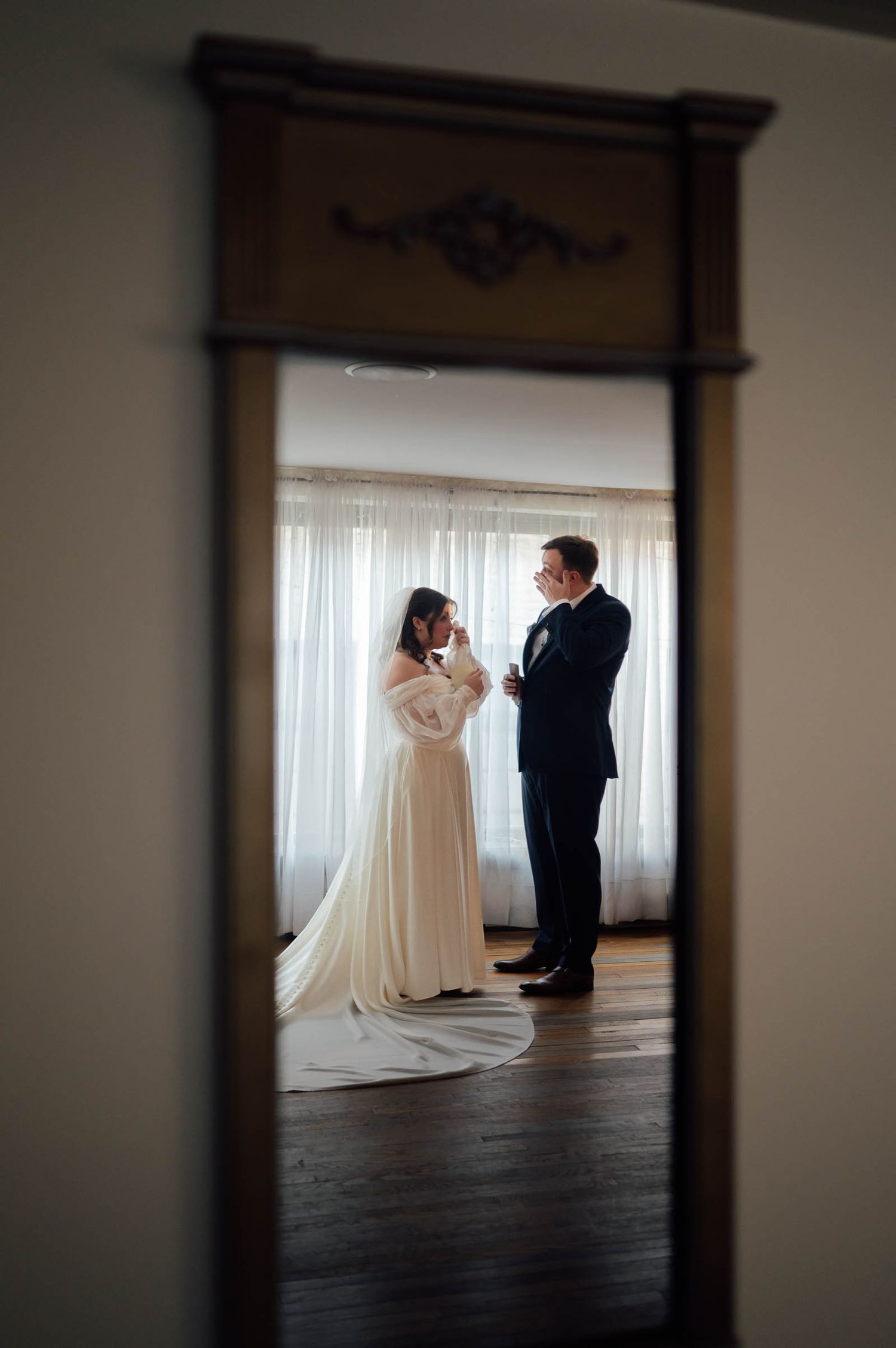 Bride in flowing white gown and groom share a private moment reflected in an antique mirror.