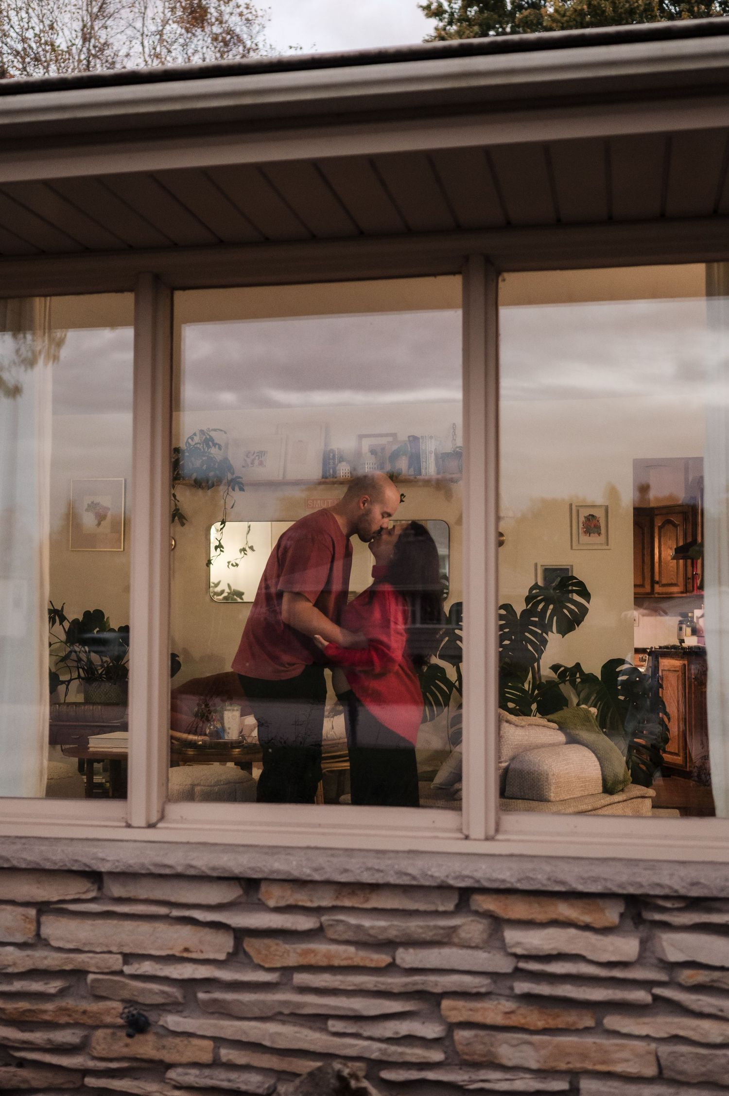 Couple sharing a kiss inside home viewed through large glass window from outside.