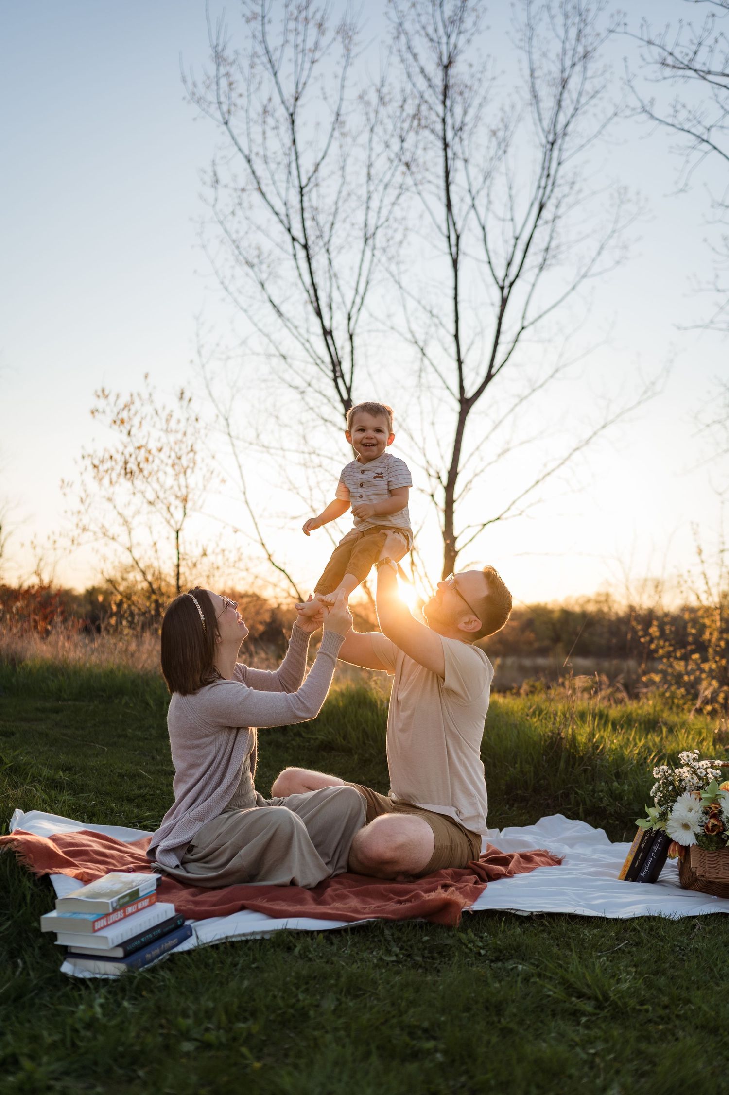 Joyful parents lift laughing baby in the air on picnic blanket during golden hour sunset outdoors.